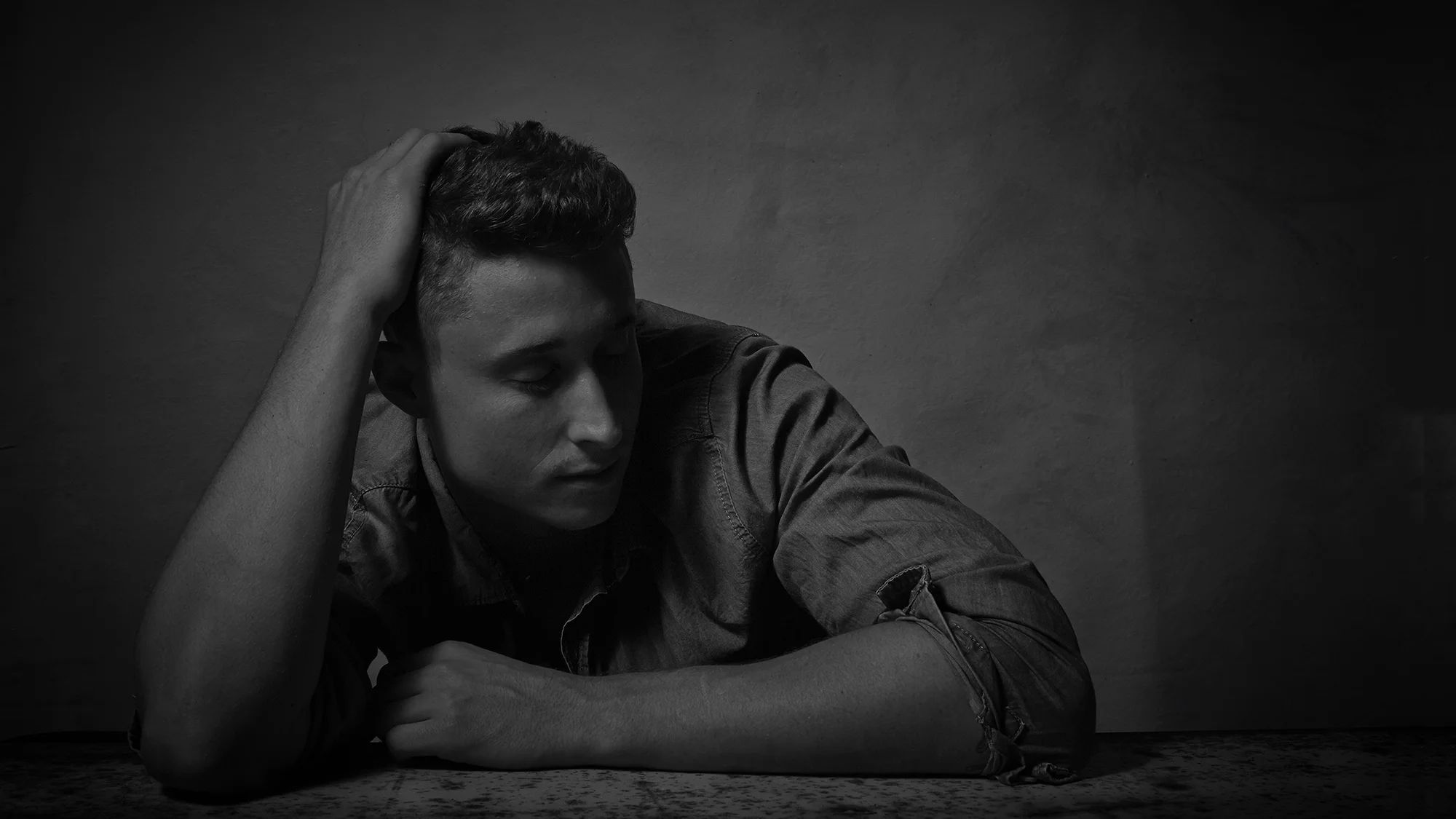 A black and white photo of a man with short hair, resting his forehead on his forearm, with one hand in his hair, against a plain wall background.