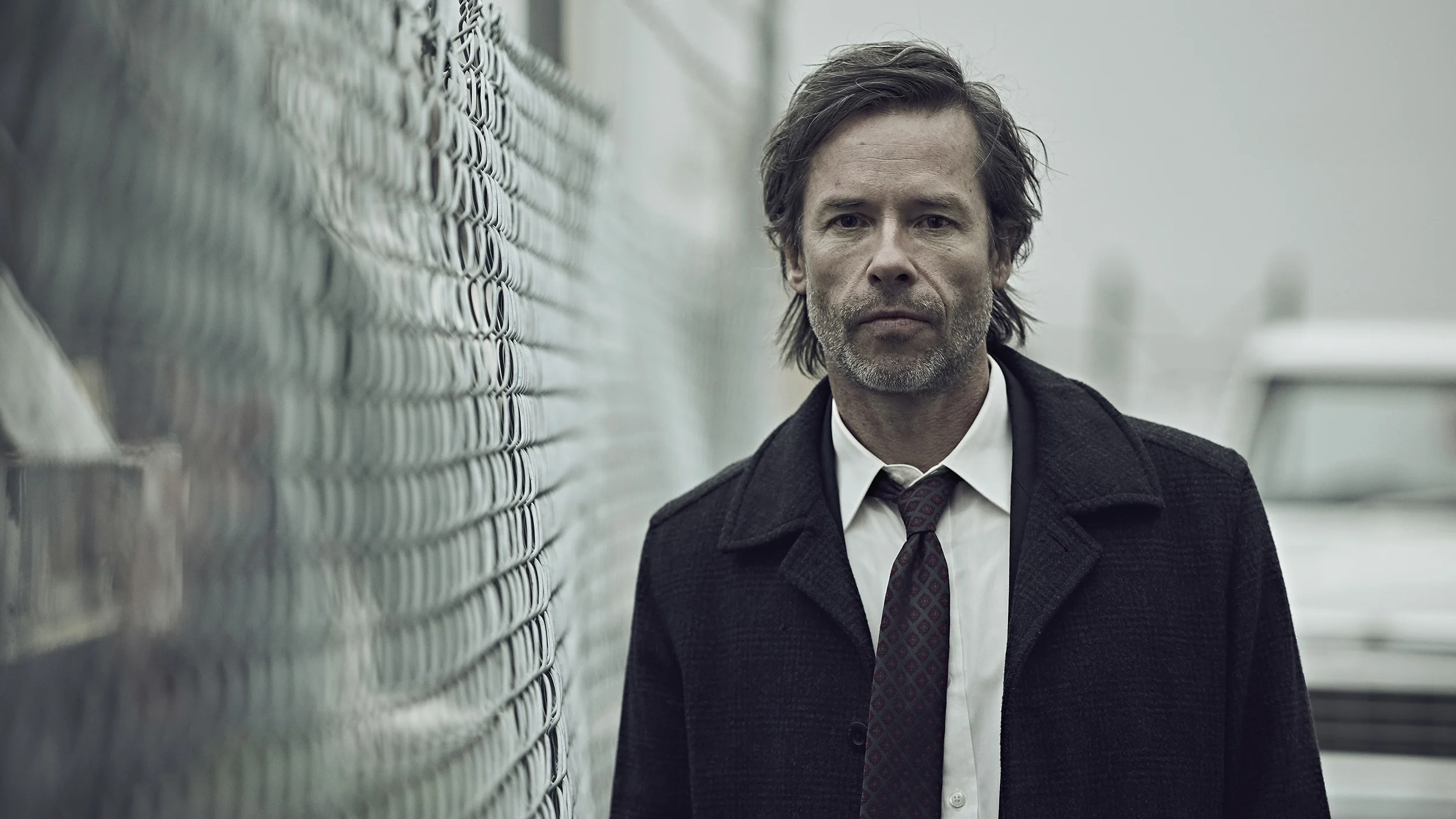 A middle-aged man with dark and gray hair, wearing a dark gray coat, white shirt, and patterned tie, stands outdoors near a chain-link fence on a foggy day.