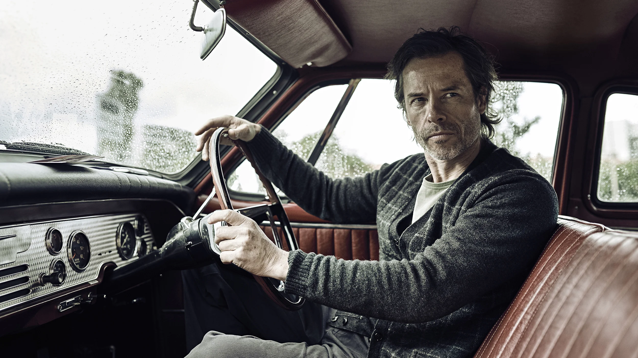 A middle-aged man with dark hair and a beard sitting in the driver's seat of a vintage car during rainy weather, looking outside the window with raindrops on the glass.