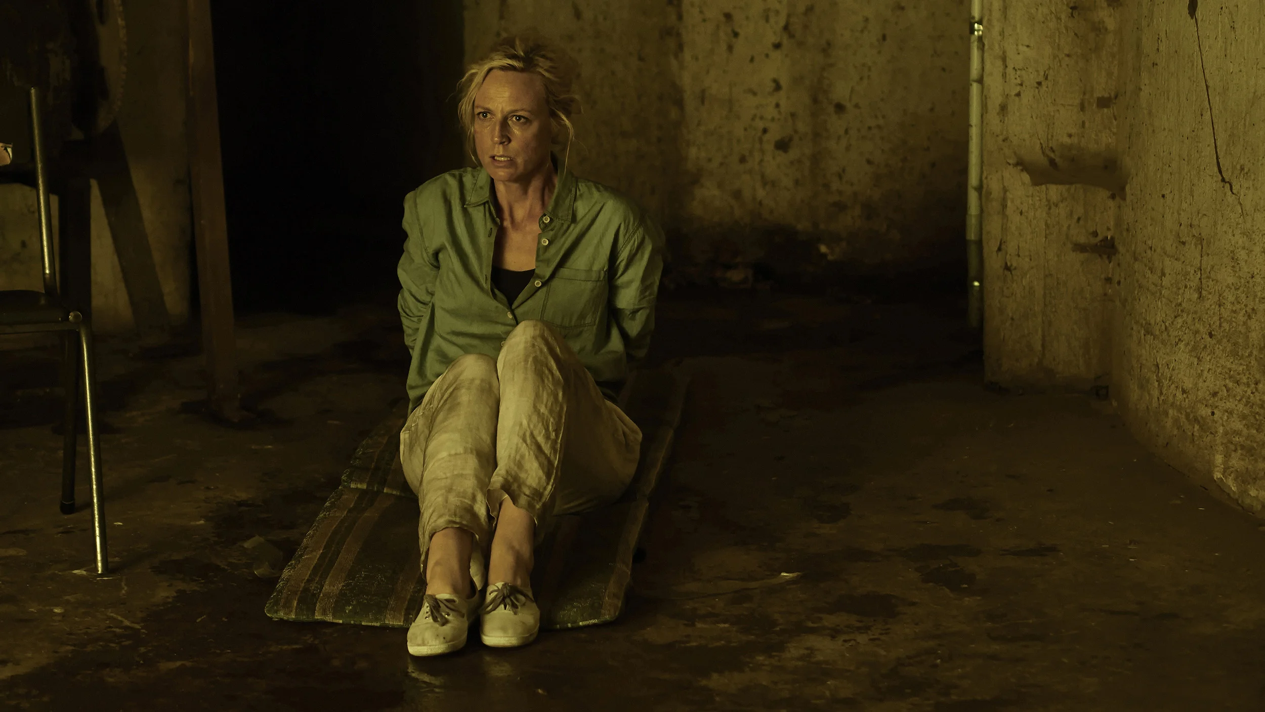 Woman sitting on a platform in a dimly lit, rough-textured basement or cellar with bare walls, wearing a green shirt and beige pants, looking distressed.