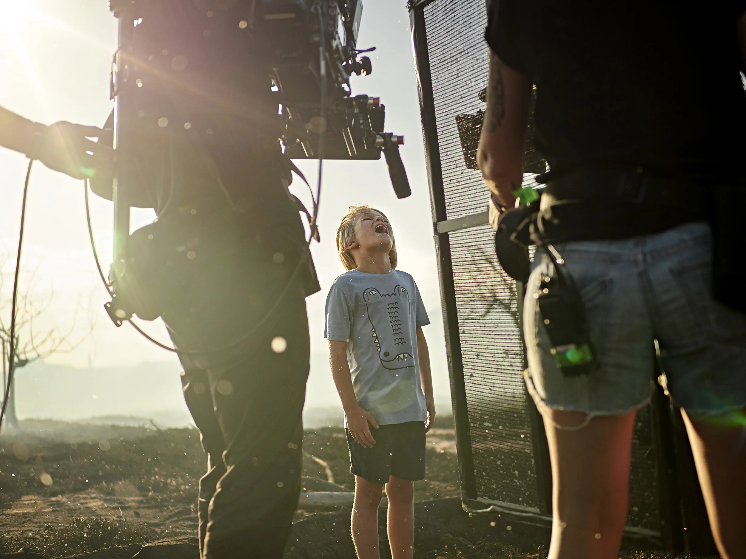 A young boy standing outdoors at sunset surrounded by adults, with a camera and filming equipment in the foreground.