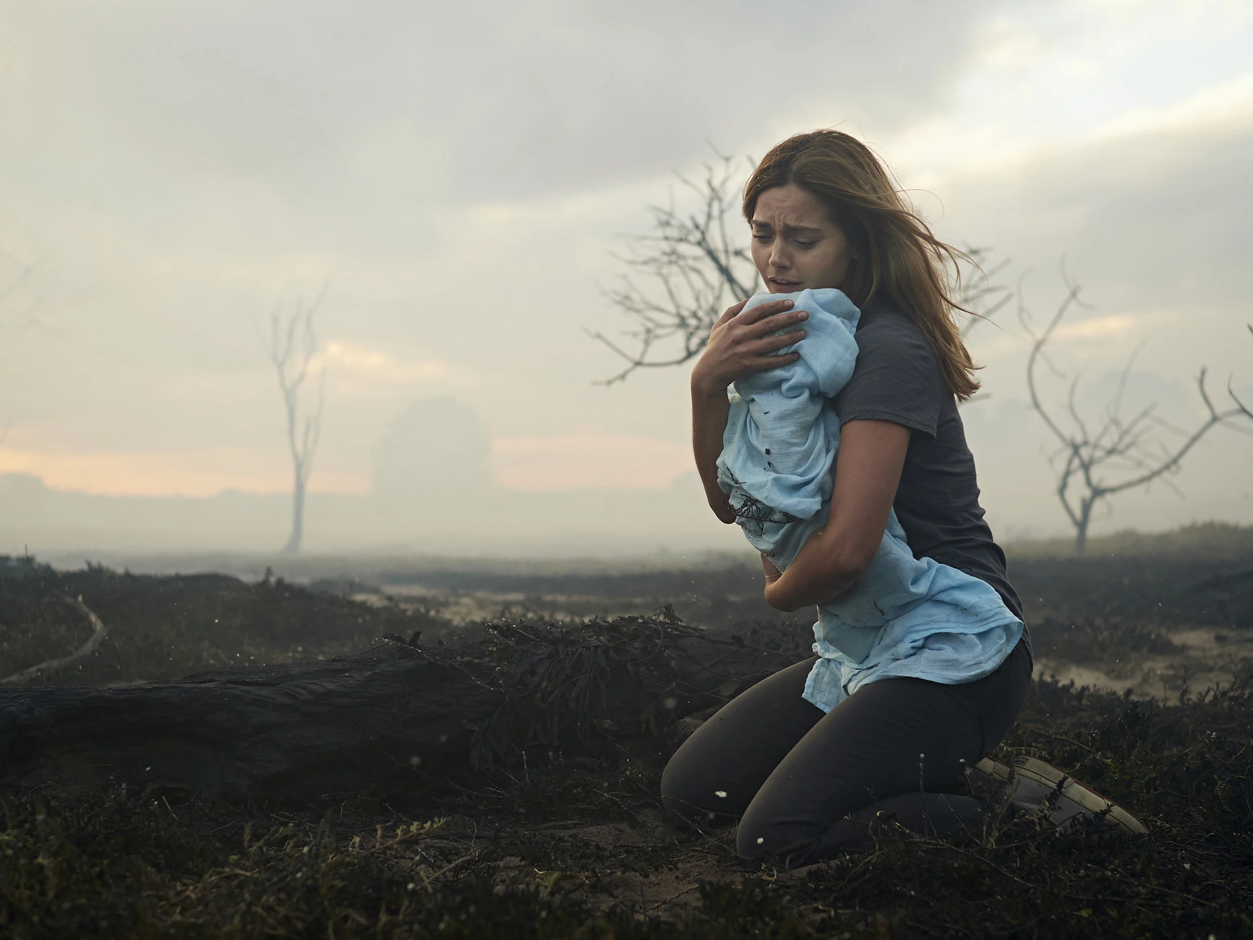 A woman kneeling on dirt and holding a crying child wrapped in a blue cloth outside during cloudy weather, with barren trees in the background.