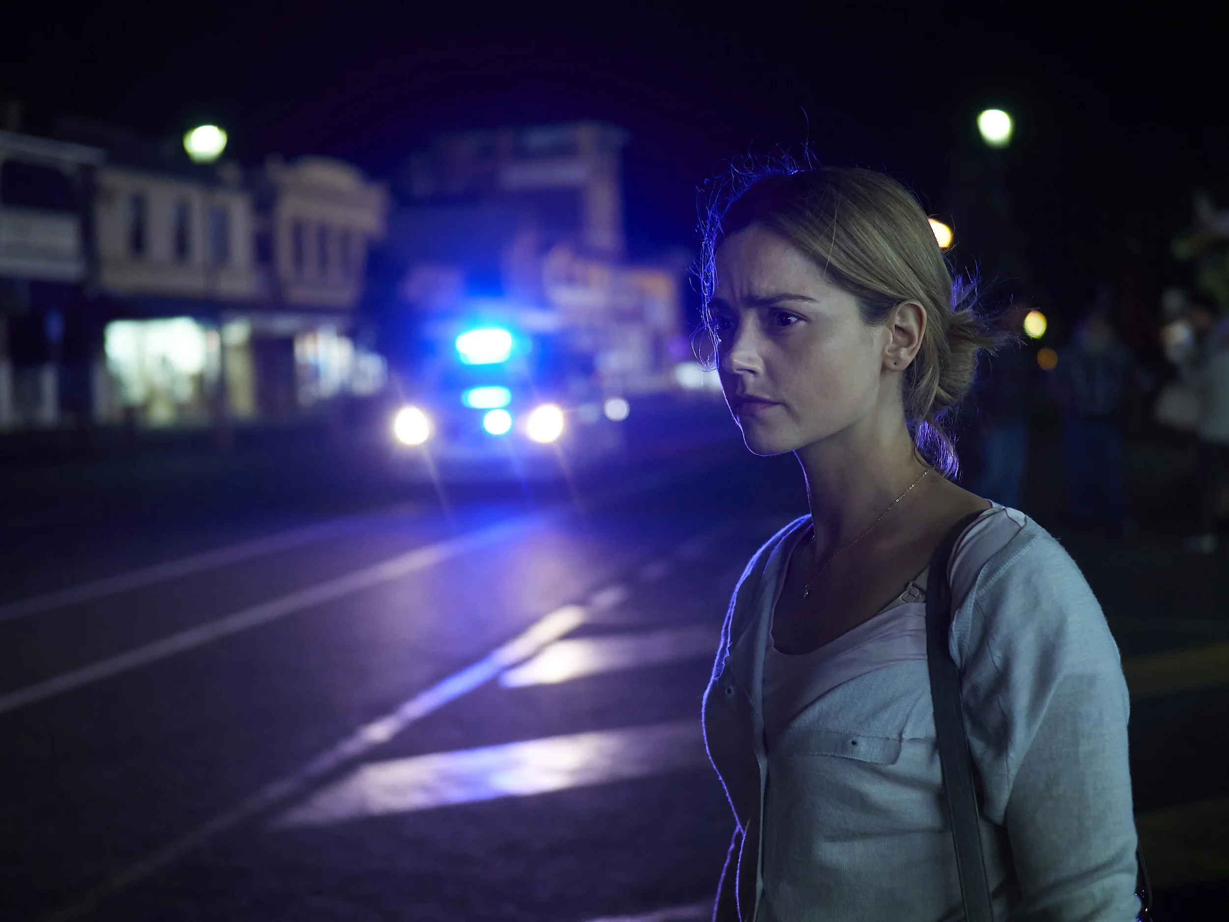 A woman with blonde hair tied back, standing on a street at night, with police car lights flashing in the background.