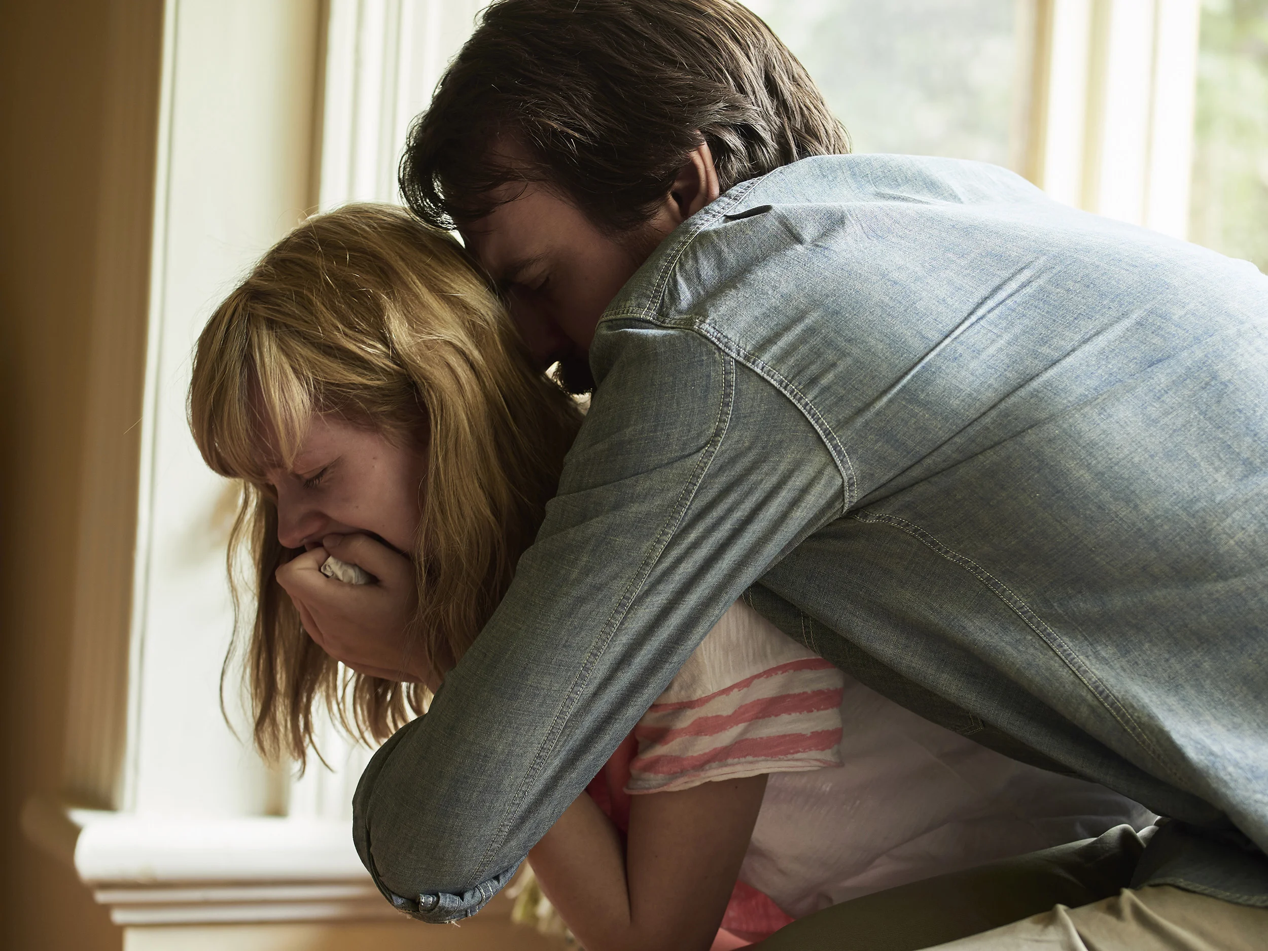 A man hugging a woman from behind, both with their heads bowed down, near a window with natural light.