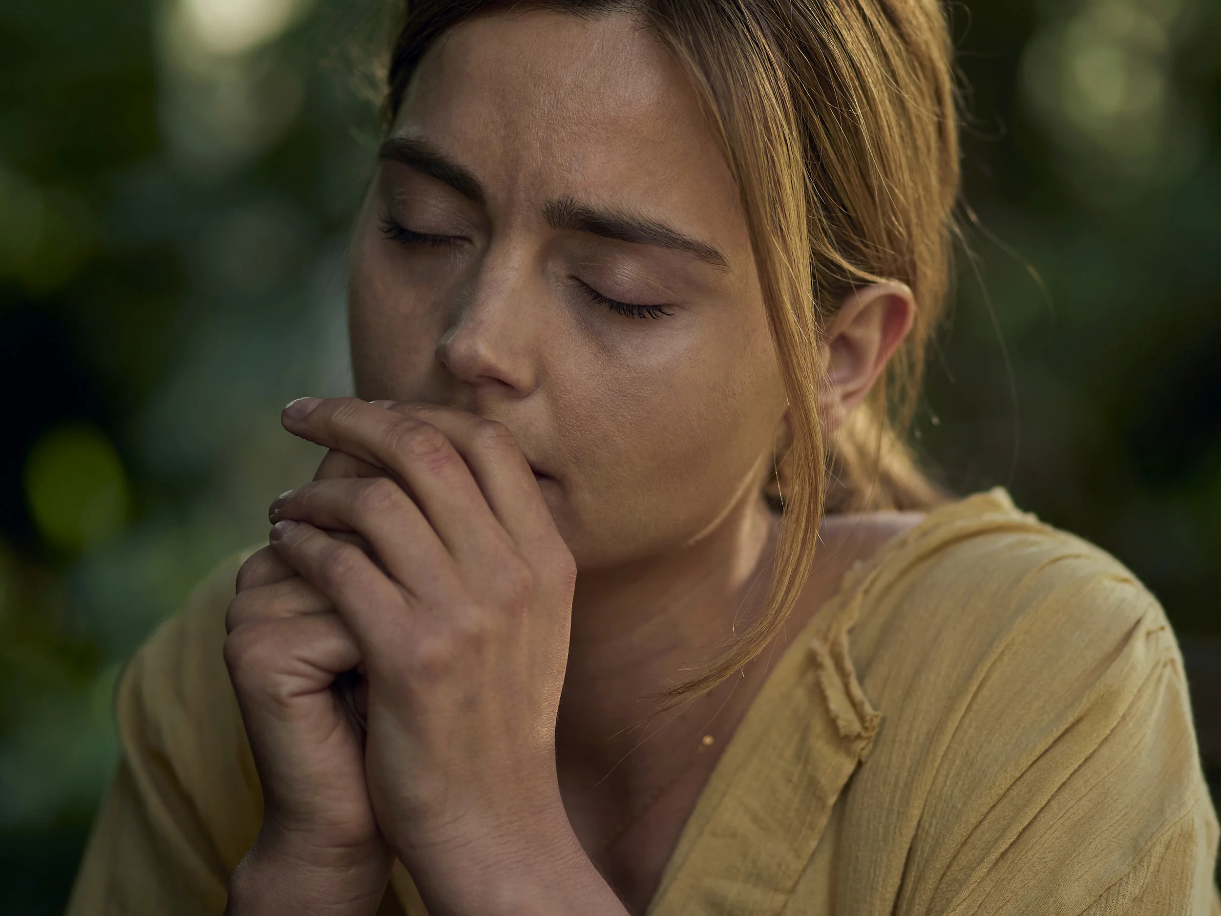A woman with closed eyes, holding her hand close to her face, outdoors with green blurred background.