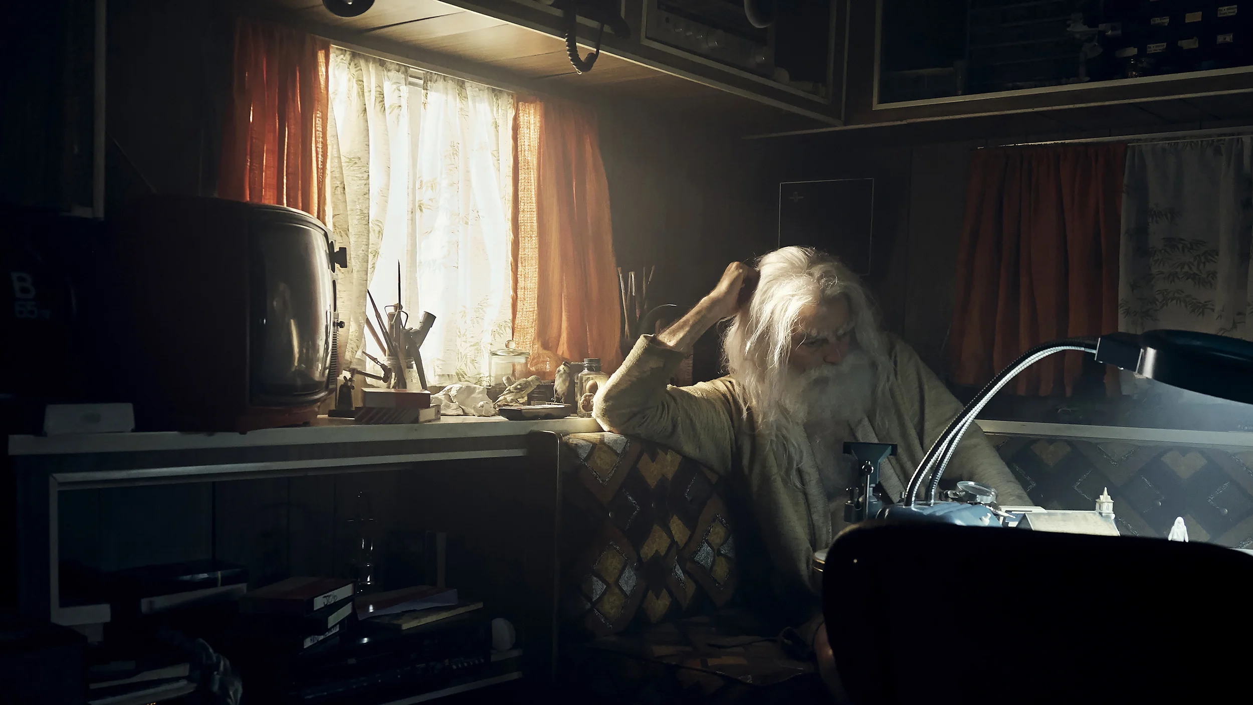 An elderly person with white hair and a beard sitting at a cluttered desk in a dimly lit room, with a window covered by curtains in the background.