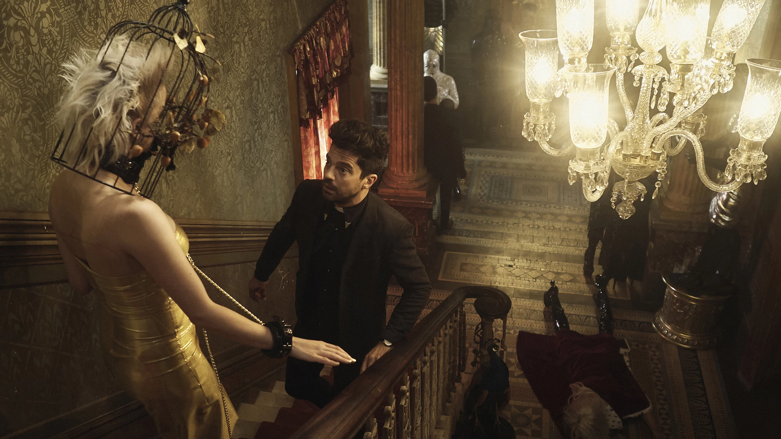 A young man dressed in black is looking up at a mannequin dressed in a gold dress and a large black hat with feathers, inside an ornate, dimly lit mansion or historic building with vintage decor and a large crystal chandelier.