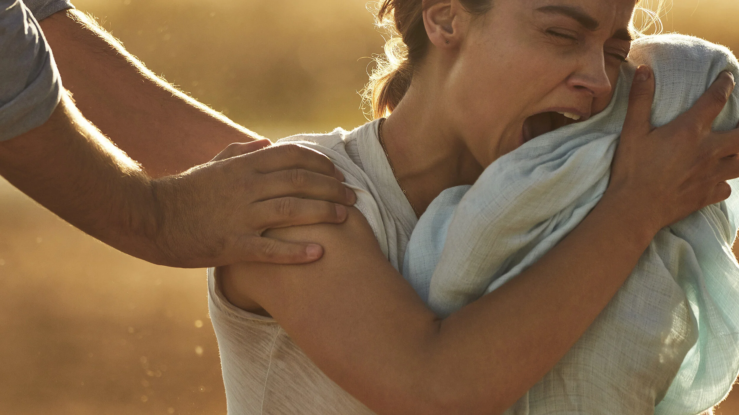 A woman being embraced from behind while yawning or singing, suggesting a moment of relaxation or breath during a workout in warm, golden light.