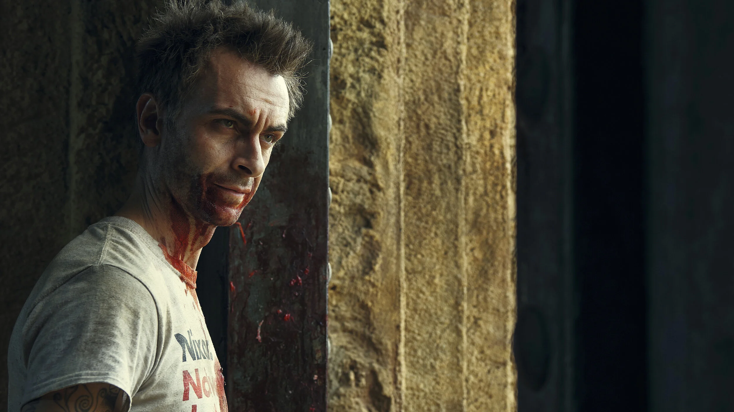 A man with blood on his face, leaning against a wall in an alleyway, looking to the side.