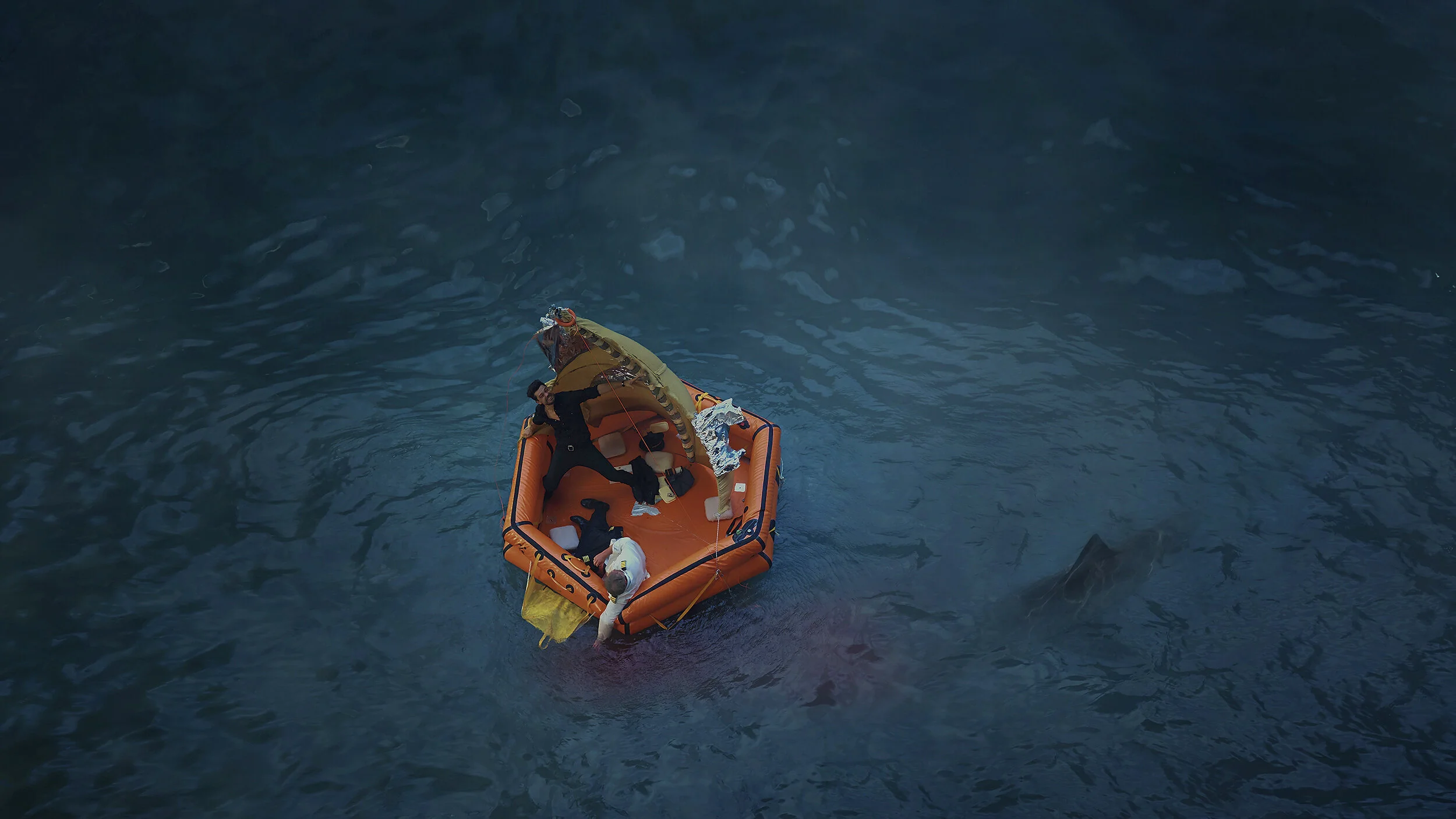 Two people in a small orange inflatable boat in dark blue water. One person is lying down, and the other is standing, holding a piece of equipment or debris.