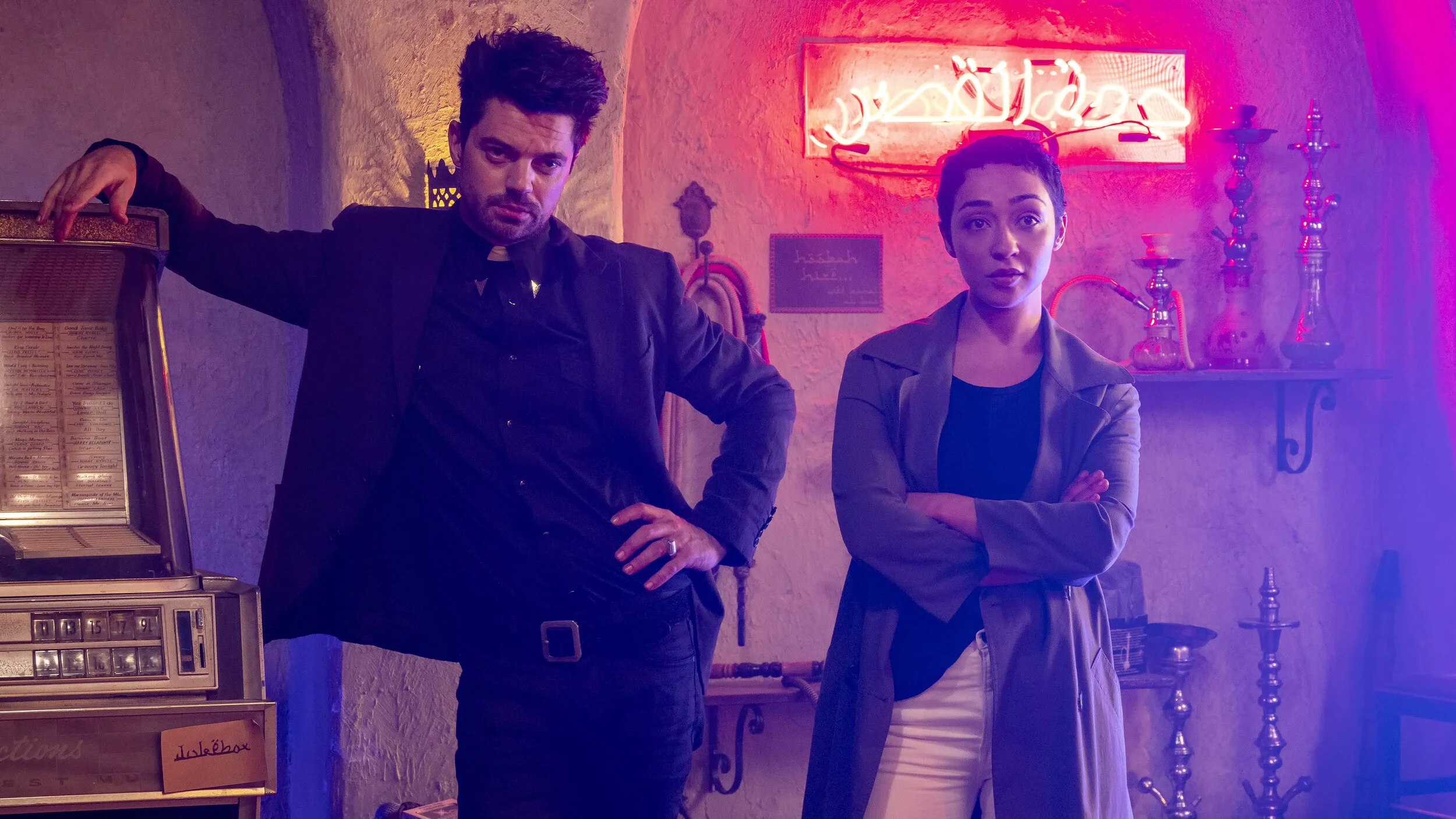 Two people standing inside a room with retro décor and hookah pipes, illuminated by pink and purple neon lights. One man in a black jacket and a woman in a gray blazer with folded arms are looking at the camera.