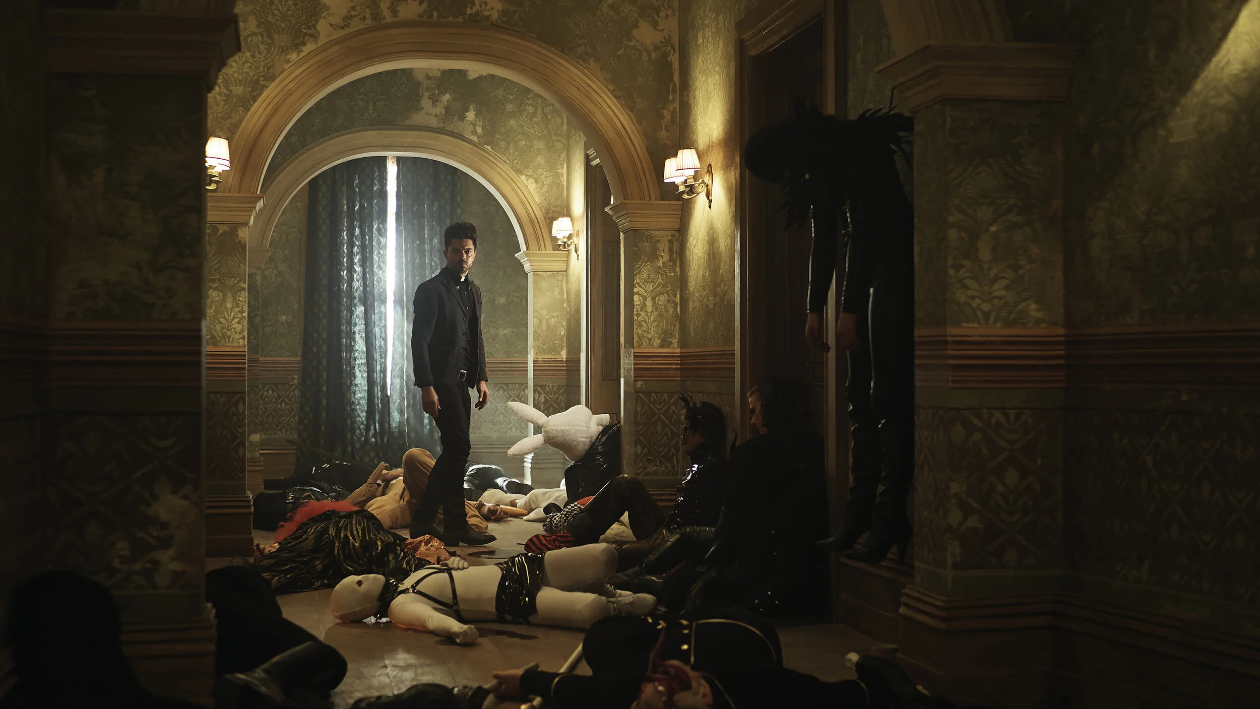 Man standing in a room with ornate wallpaper, multiple people lying on the floor around him, some in costumes or masks, in a dimly lit setting with wall-mounted lamps.