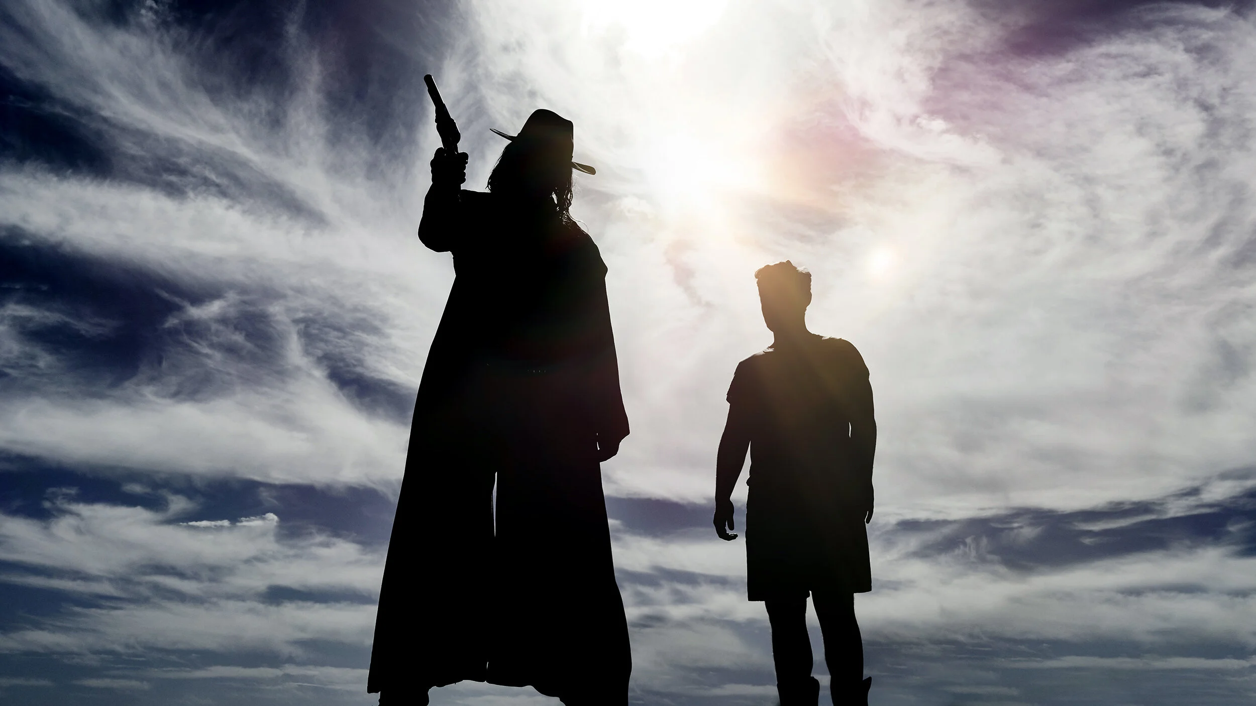 Silhouettes of two people, one woman with a hat, holding a knife and a handgun, and a man standing nearby, against a cloudy sky with the sun. TV series Preacher