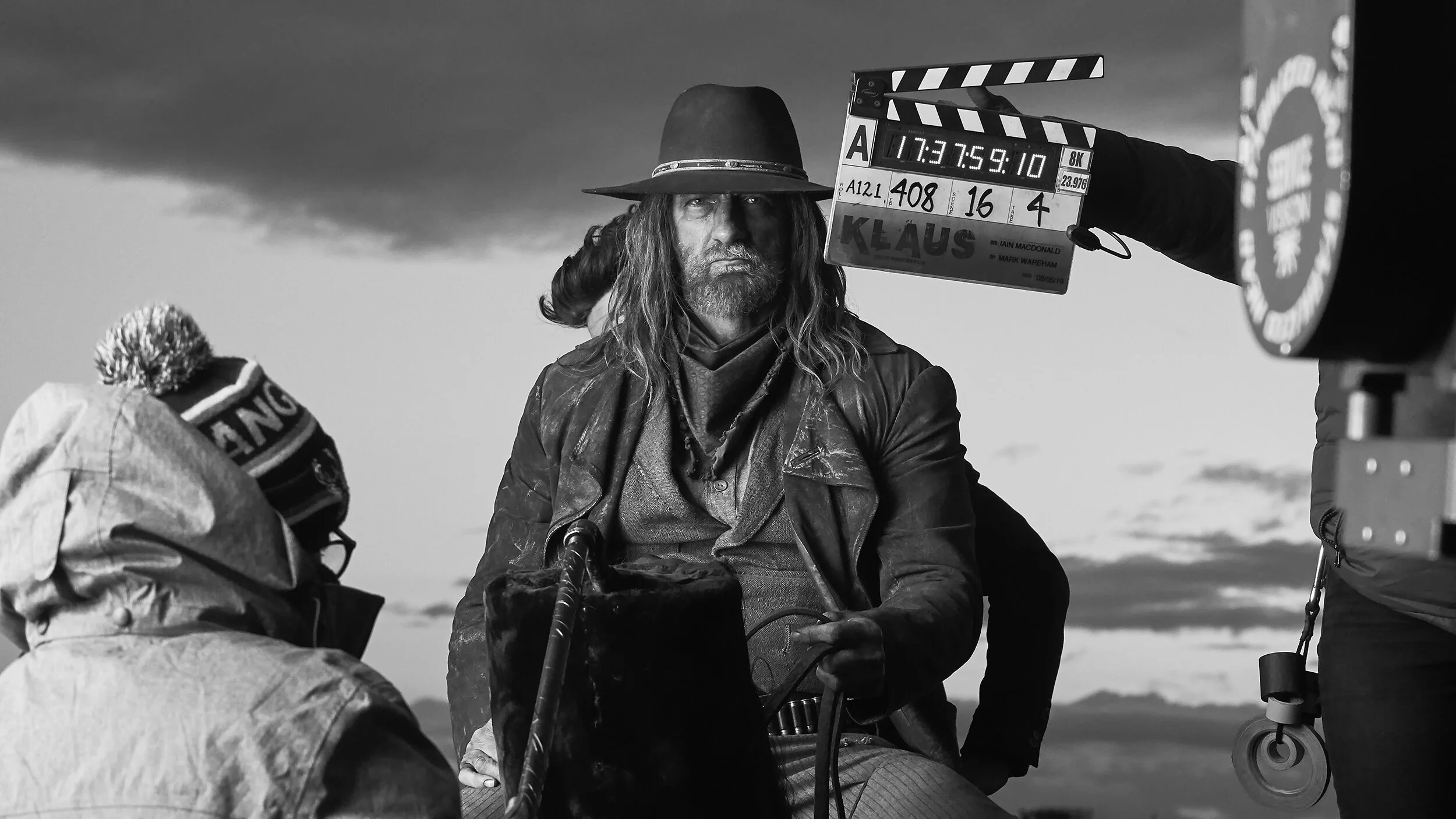 Black and white photo of a man with long hair and beard, wearing a wide-brimmed hat, sitting outdoors next to a woman with a pom-pom hat, while a clapperboard is held above them on a film set.