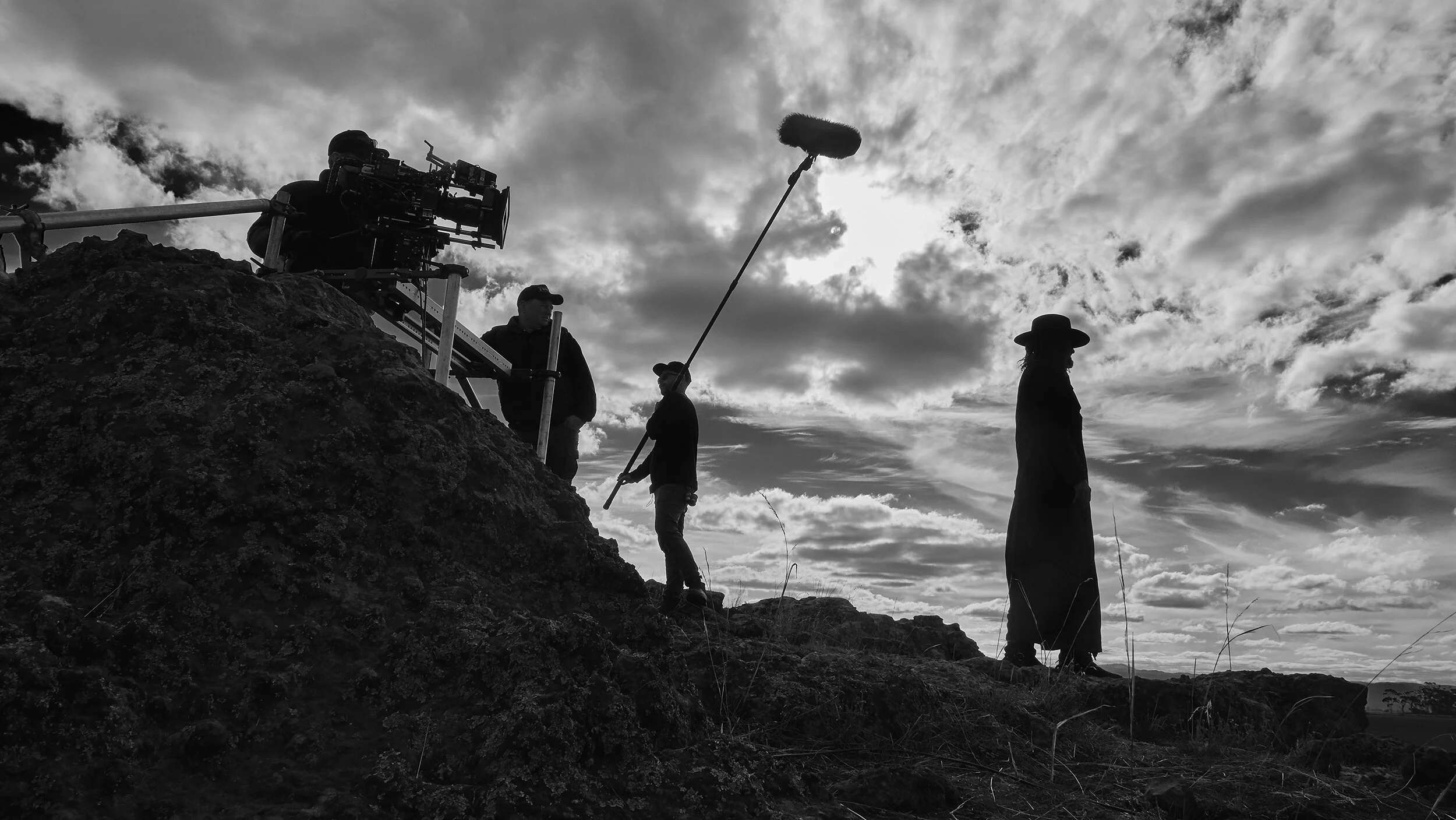 Silhouettes of crew members on a rocky terrain filming a scene, with clouds in the sky and a person holding a boom microphone in the foreground.