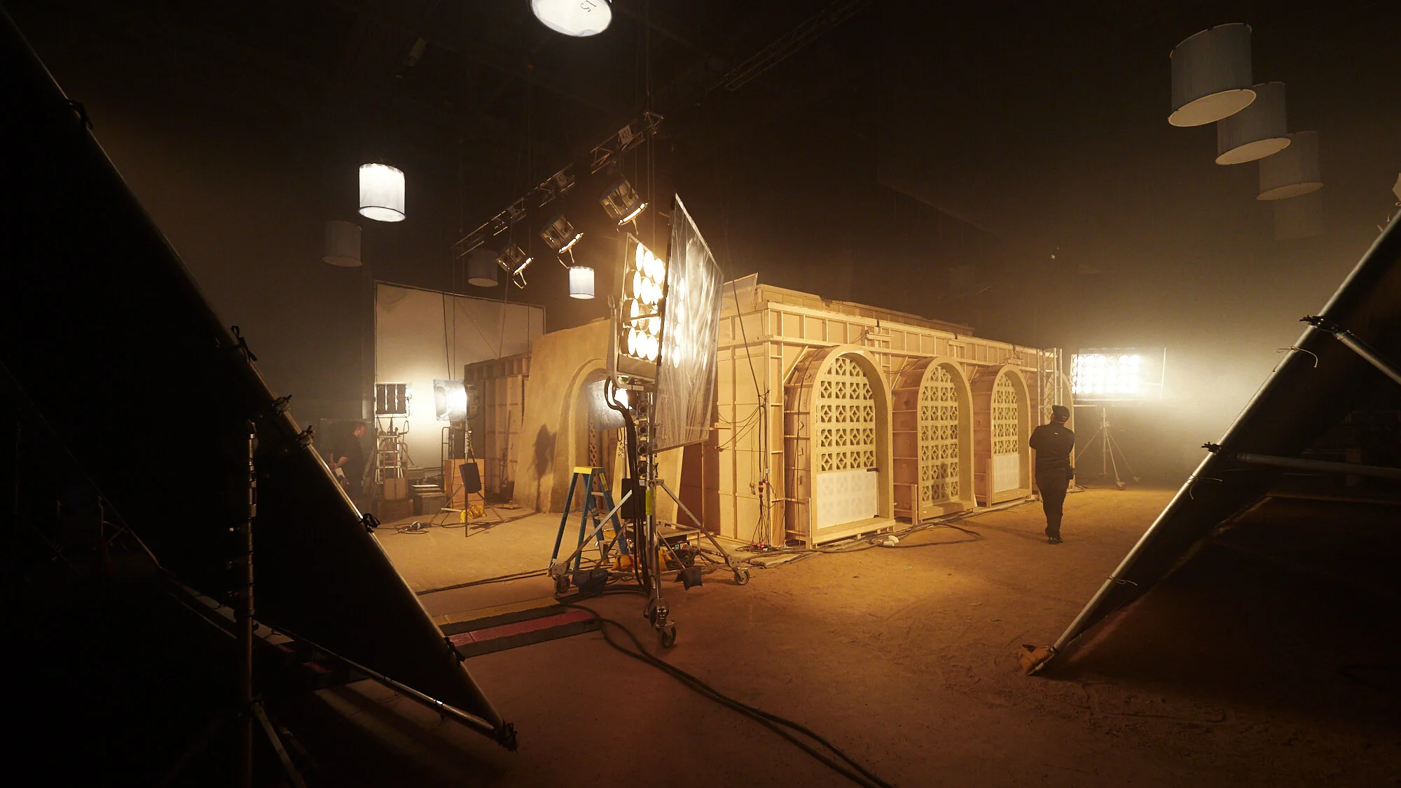 A behind-the-scenes view of a film or television set under construction with lighting and camera equipment, and a person walking in front of a wooden set design.