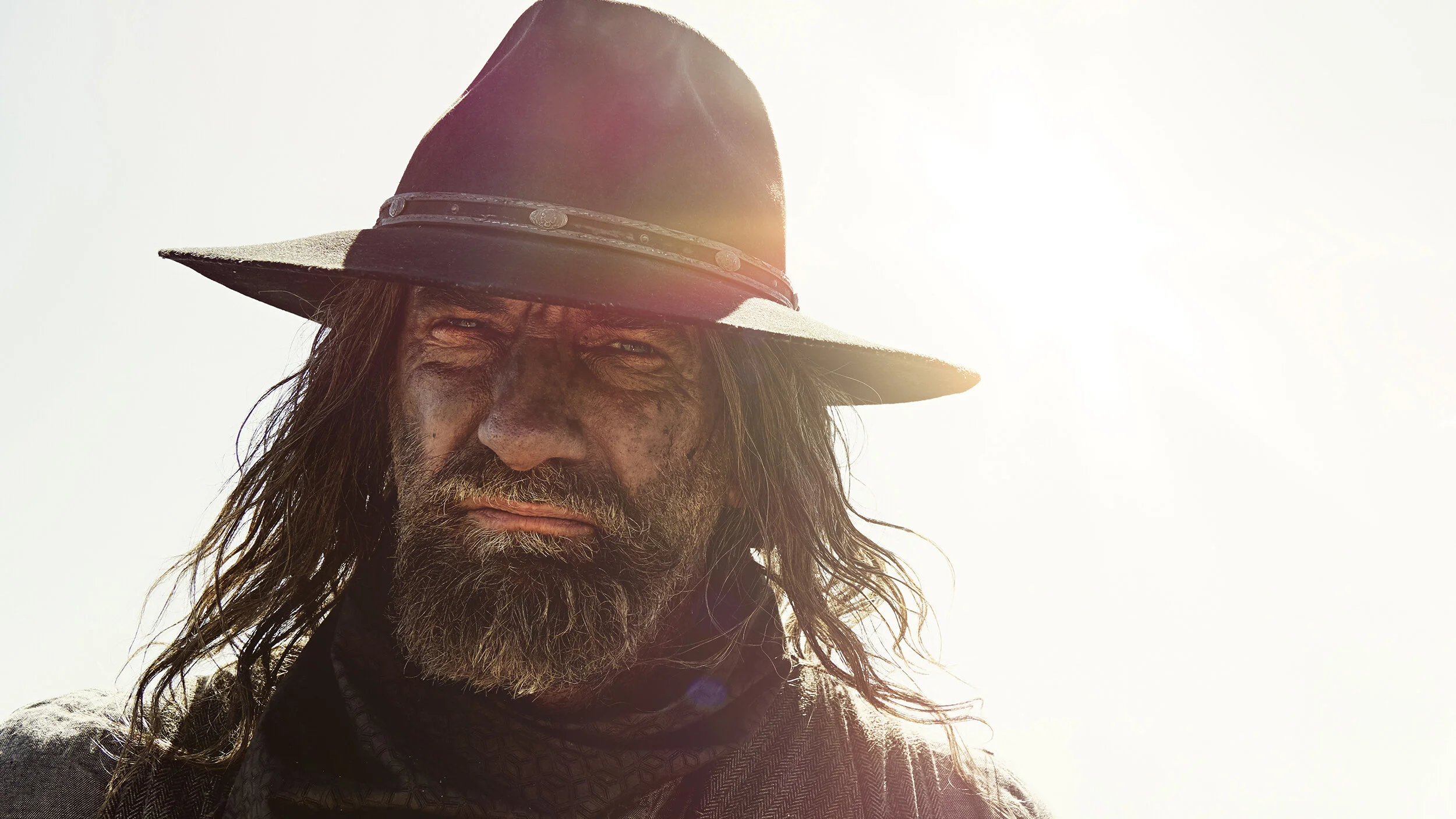 Close-up of a rugged man with long hair, beard, and mustache wearing a wide-brimmed hat with sunlight in the background.