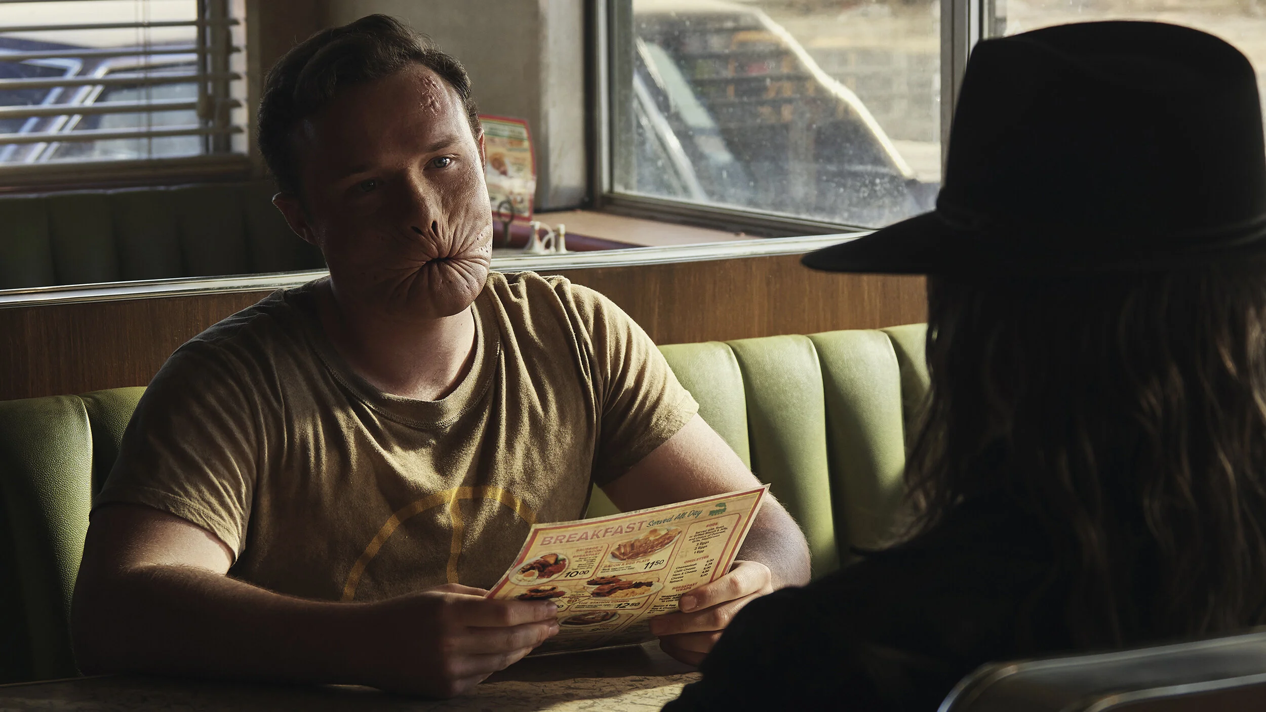 A young man is sitting in a diner booth across from a young woman wearing a black hat. He is holding a menu and looks at her with a humorous, distorted face.  TV series Preacher