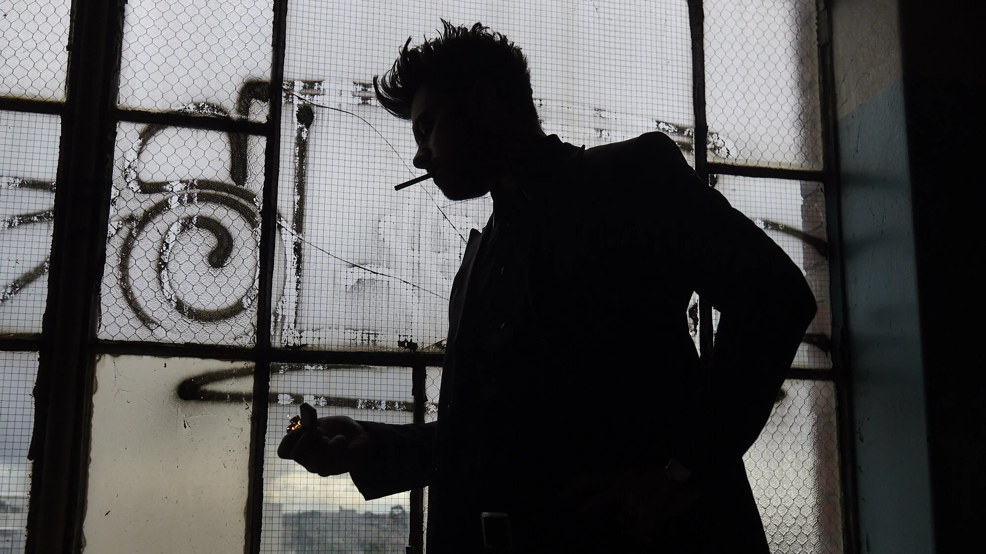 Silhouette of a person with styled hair holding a smartphone and a cigarette, standing in front of a large window with broken glass and graffiti on the outside.