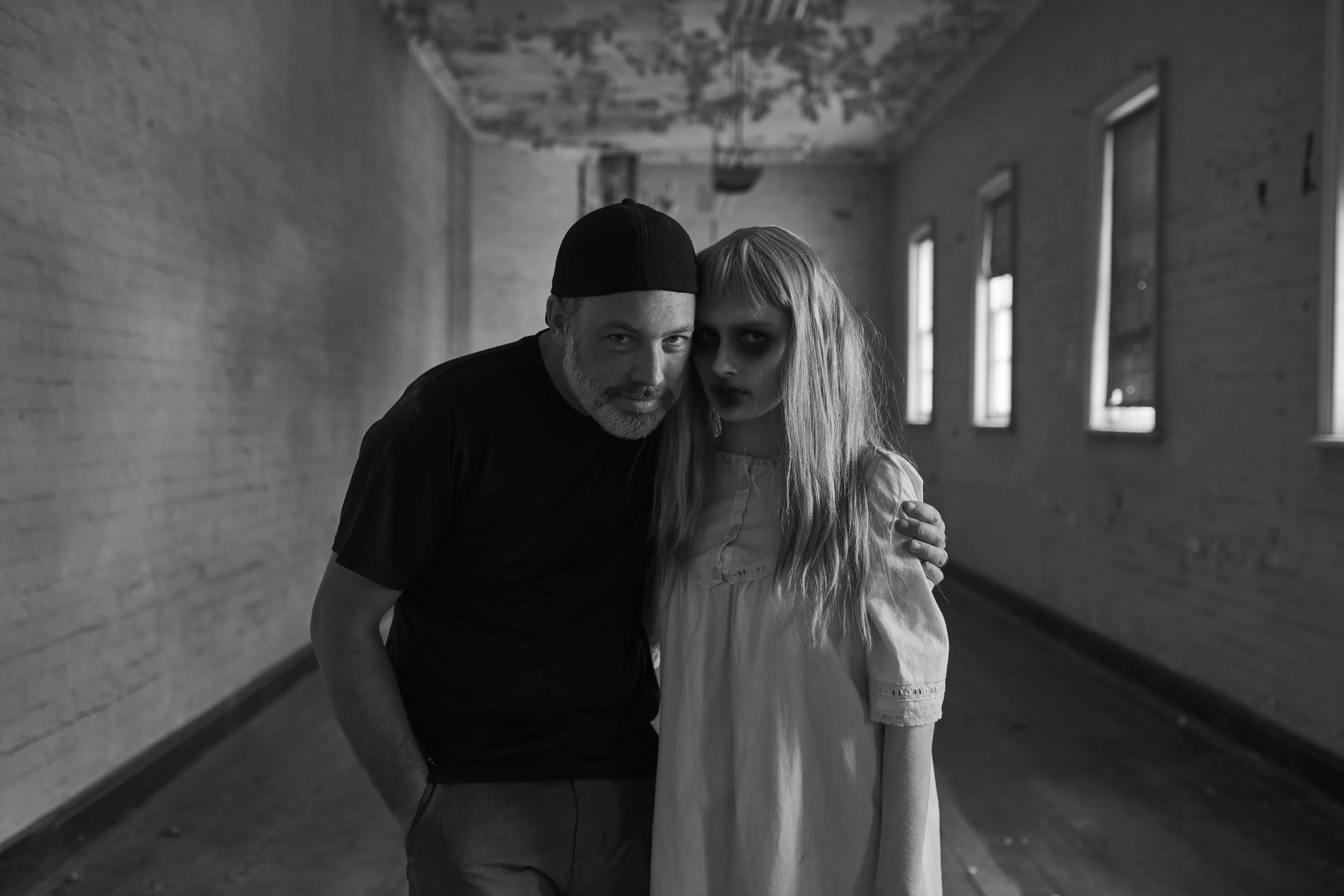 A black and white photo of a man and young girl standing close together in a long, empty hallway with windows. The man has a beard, wears a black beanie, and a dark t-shirt. The girl has long, wavy hair, dark makeup around her eyes, and is wearing a 