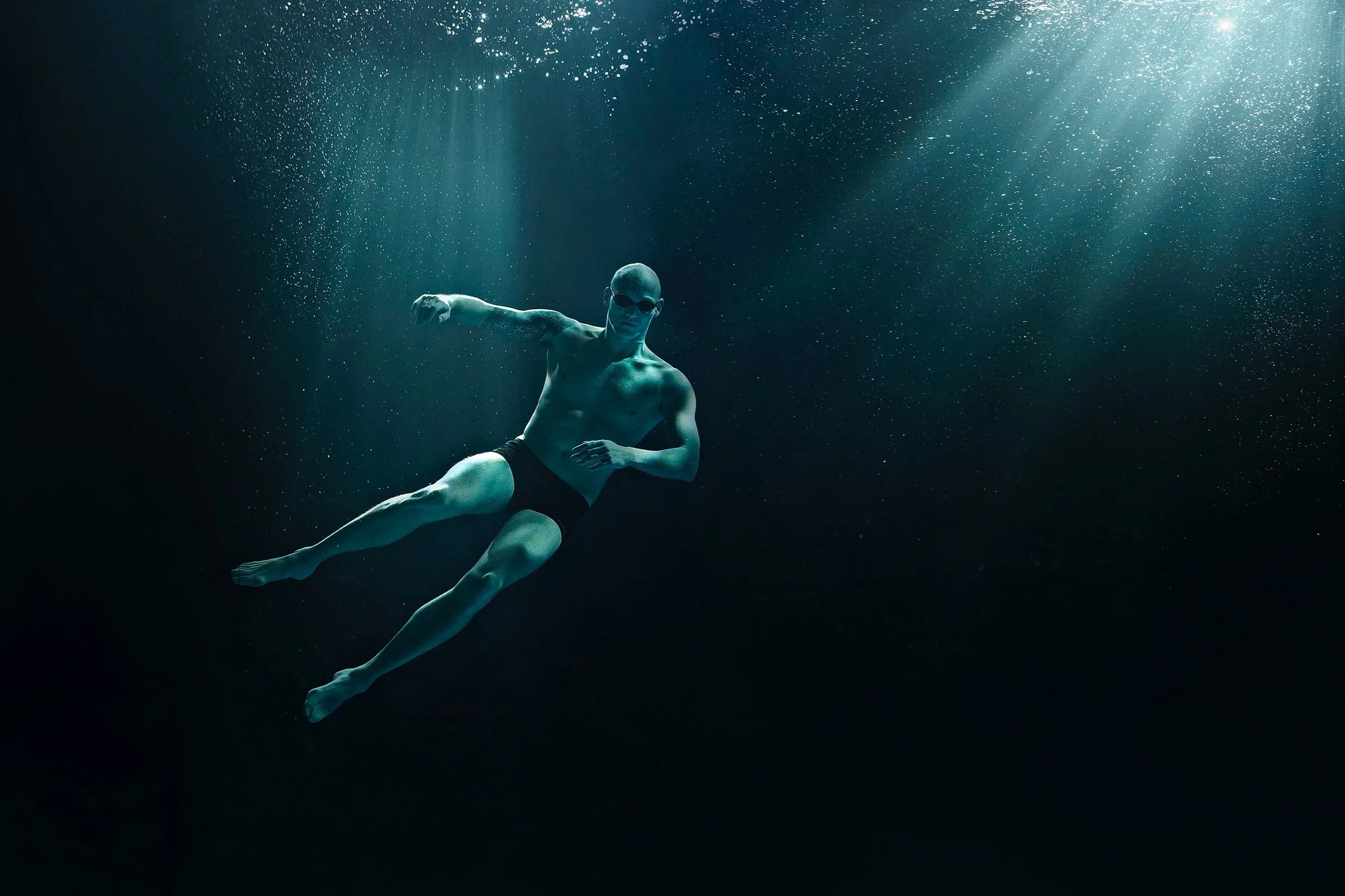 A man swimming underwater, wearing goggles, surrounded by bubbles and with light rays shining down.