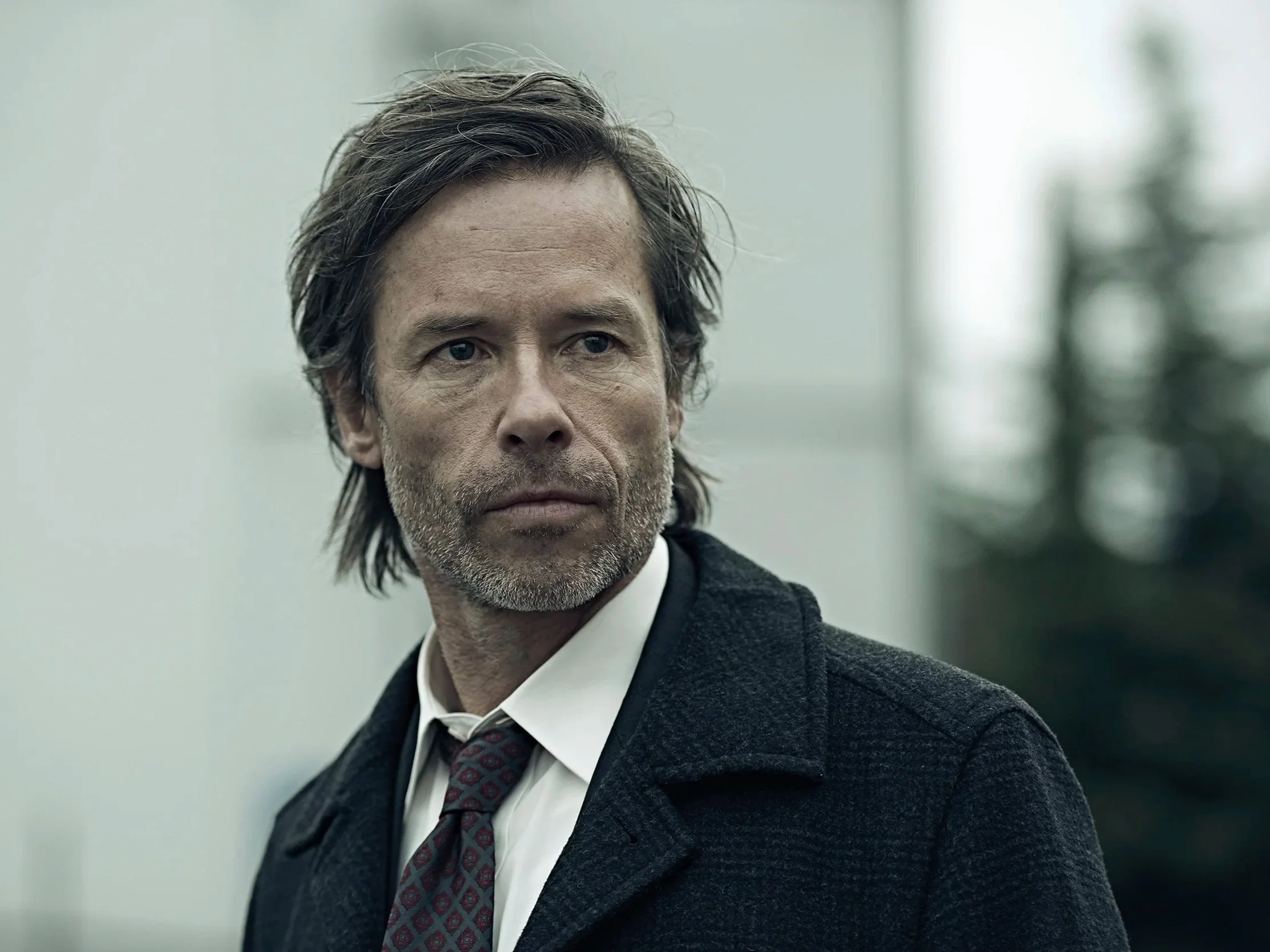 Close-up of a middle-aged man with slightly messy brown hair, wearing a dark coat, white shirt, and a patterned tie, standing outdoors with a blurred background.