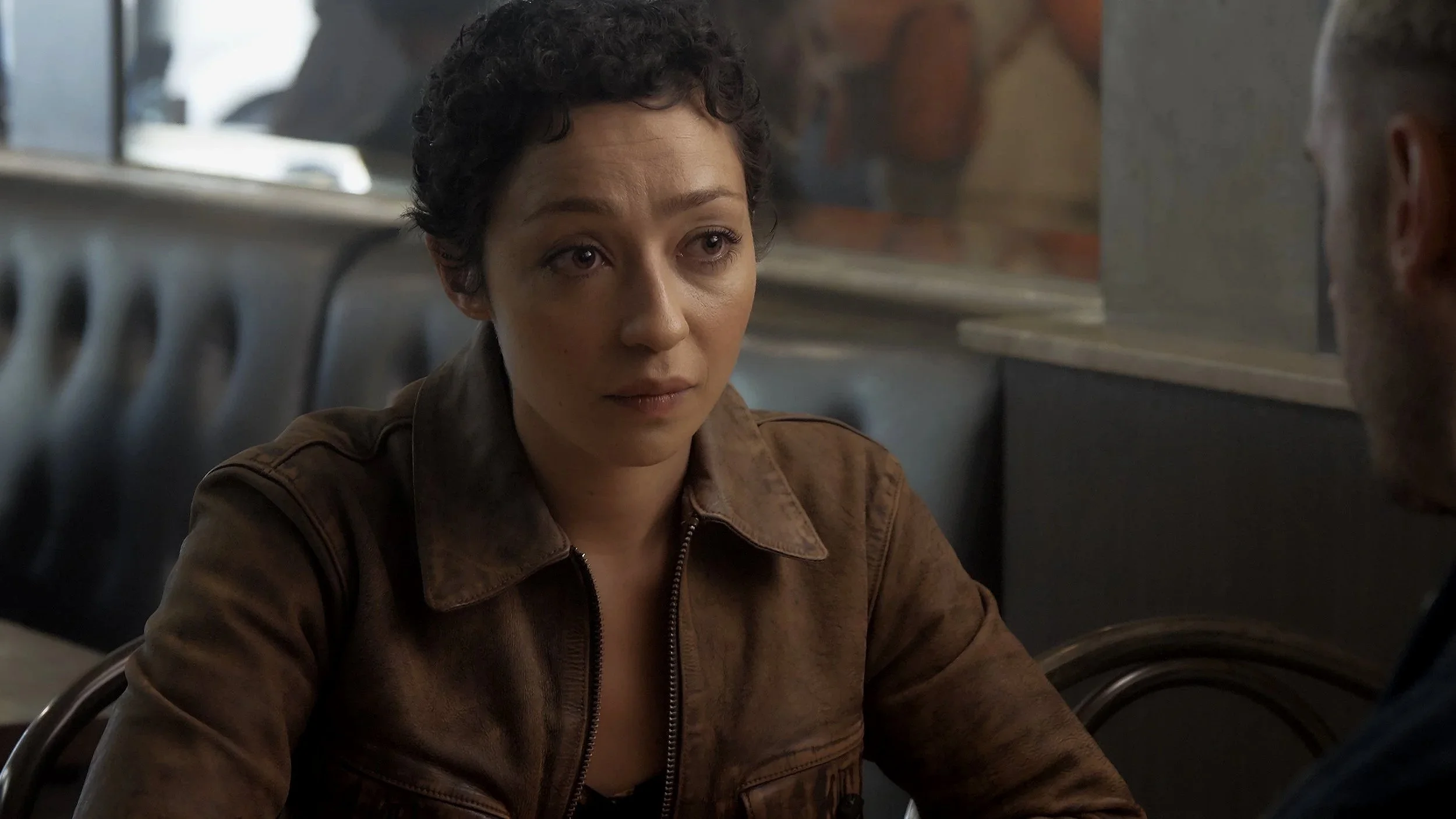 A woman with short curly hair wearing a brown jacket, sitting inside a diner or casual restaurant, looking at someone who is partially visible.