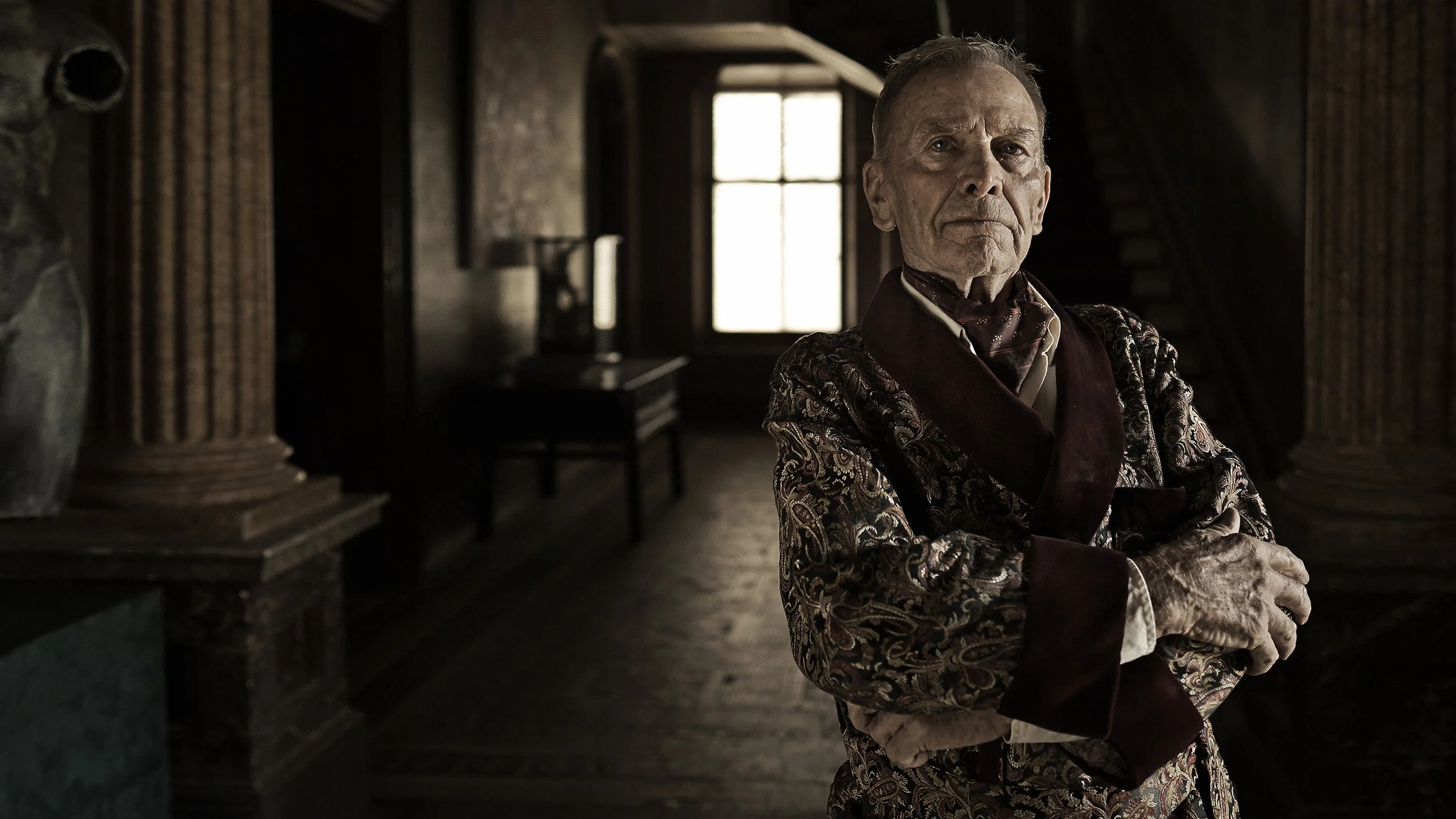 An older man with a stern expression, wearing an ornate patterned jacket and a cravat, standing inside a dimly lit room with antique decor and large windows in the background.