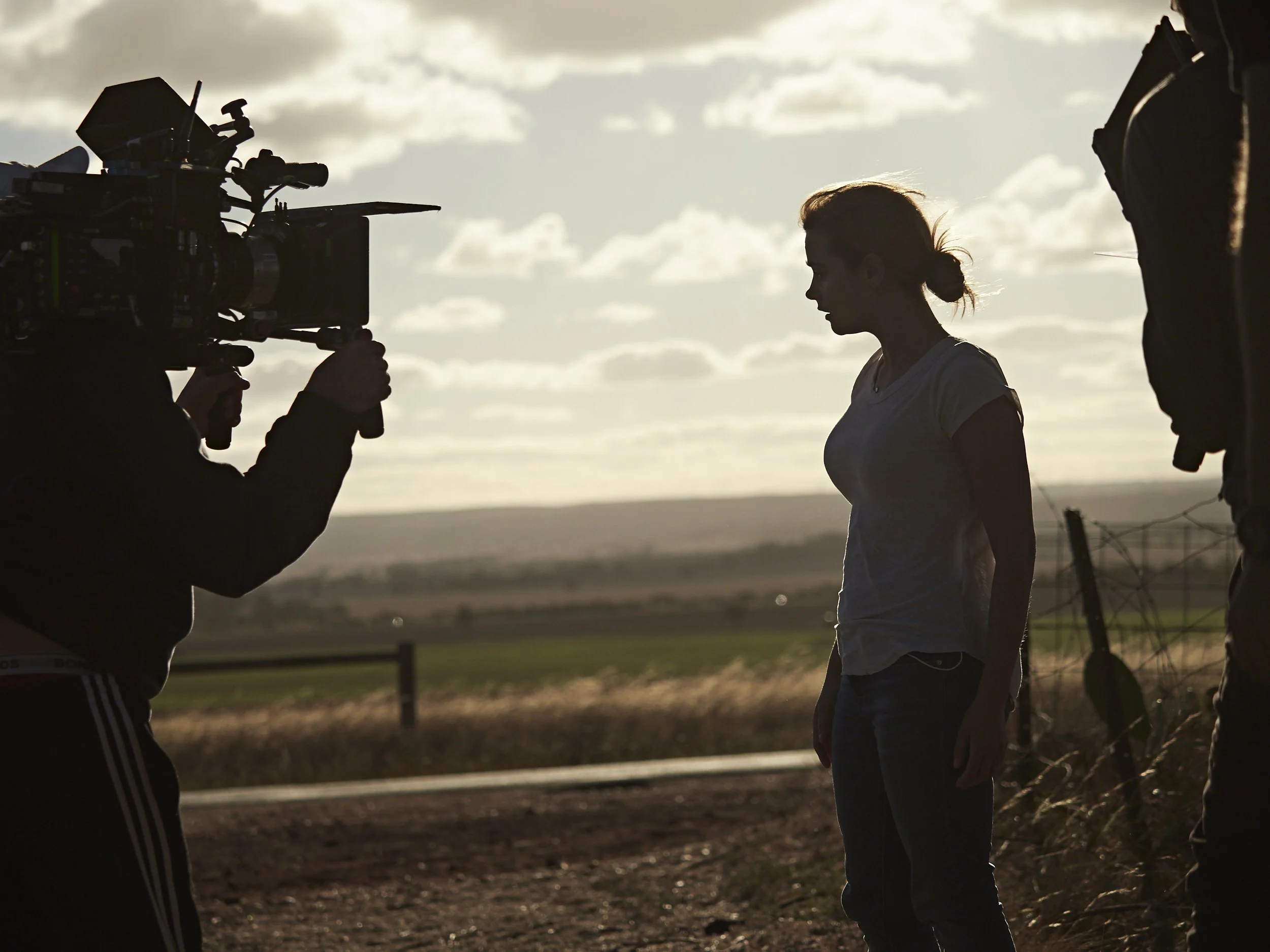 A woman with red hair in a ponytail in silhouette during a photo shoot outdoors at sunset, surrounded by two camera operators holding video cameras on tripods.