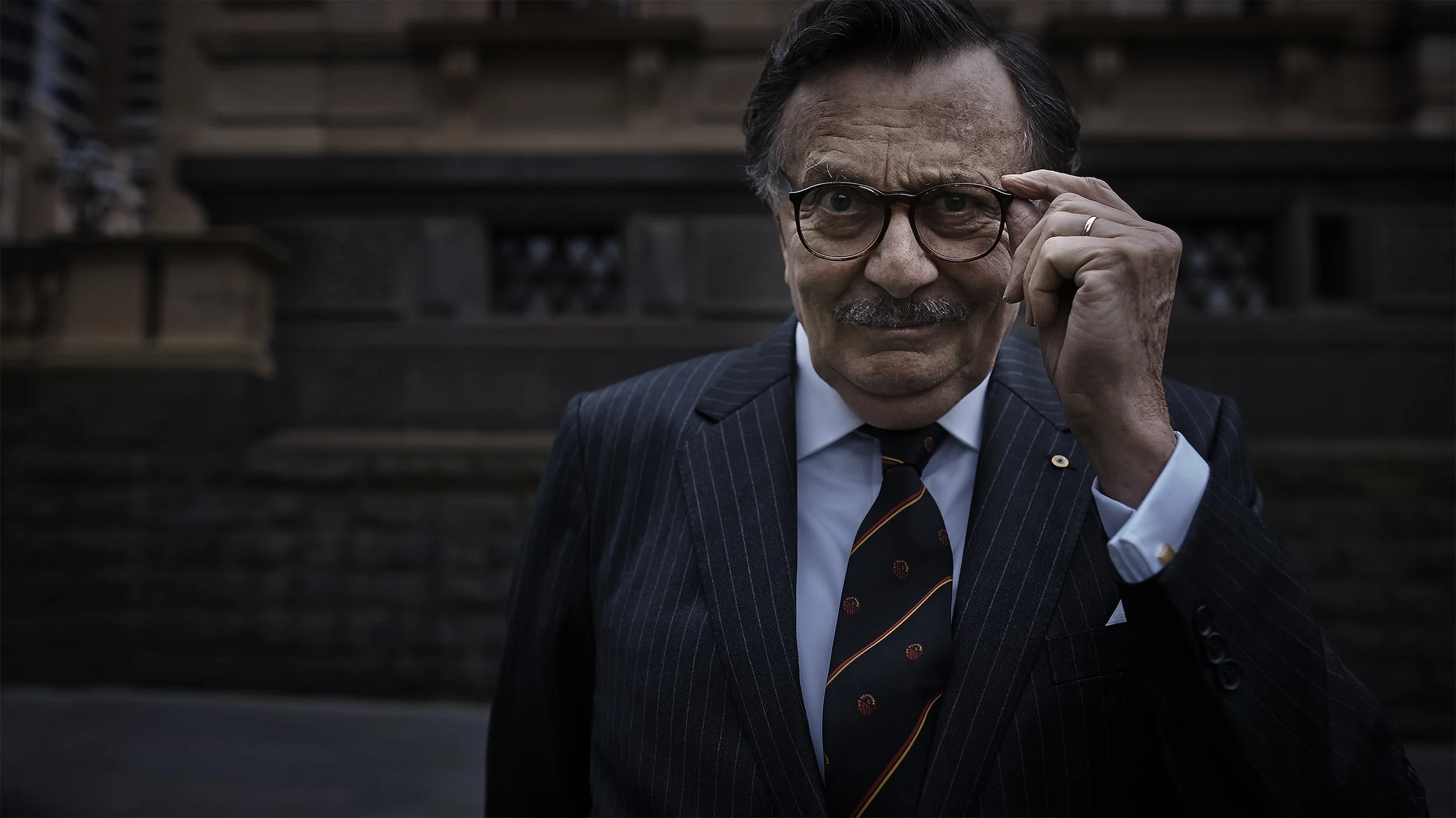A man adjusting his glasses while wearing a pinstripe suit and a tie, standing outdoors in front of a dark wooden background.