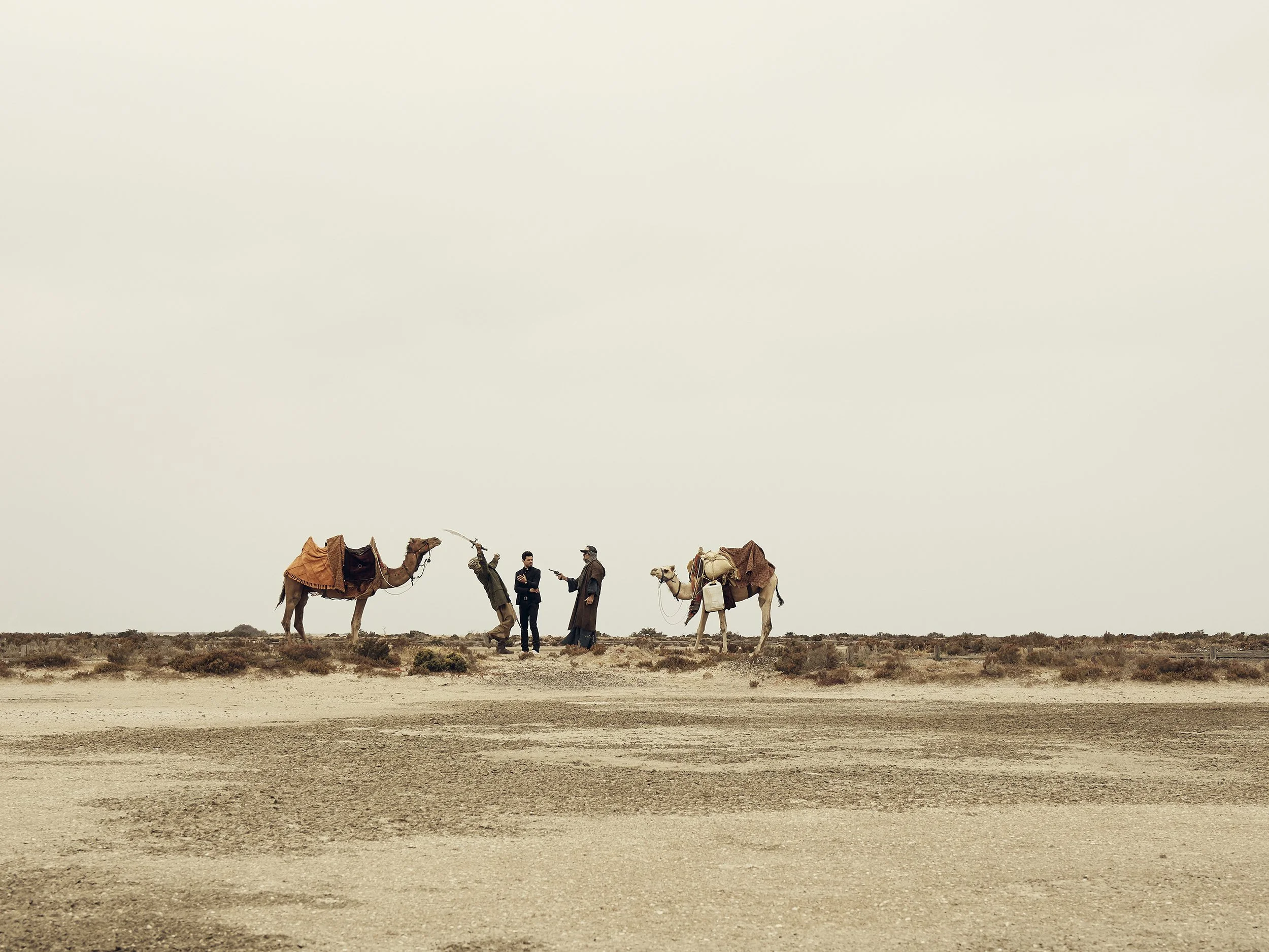 Two men talking in a desert with two camels behind them, one with a poncho and the other with a blanket, and a plain, overcast sky.