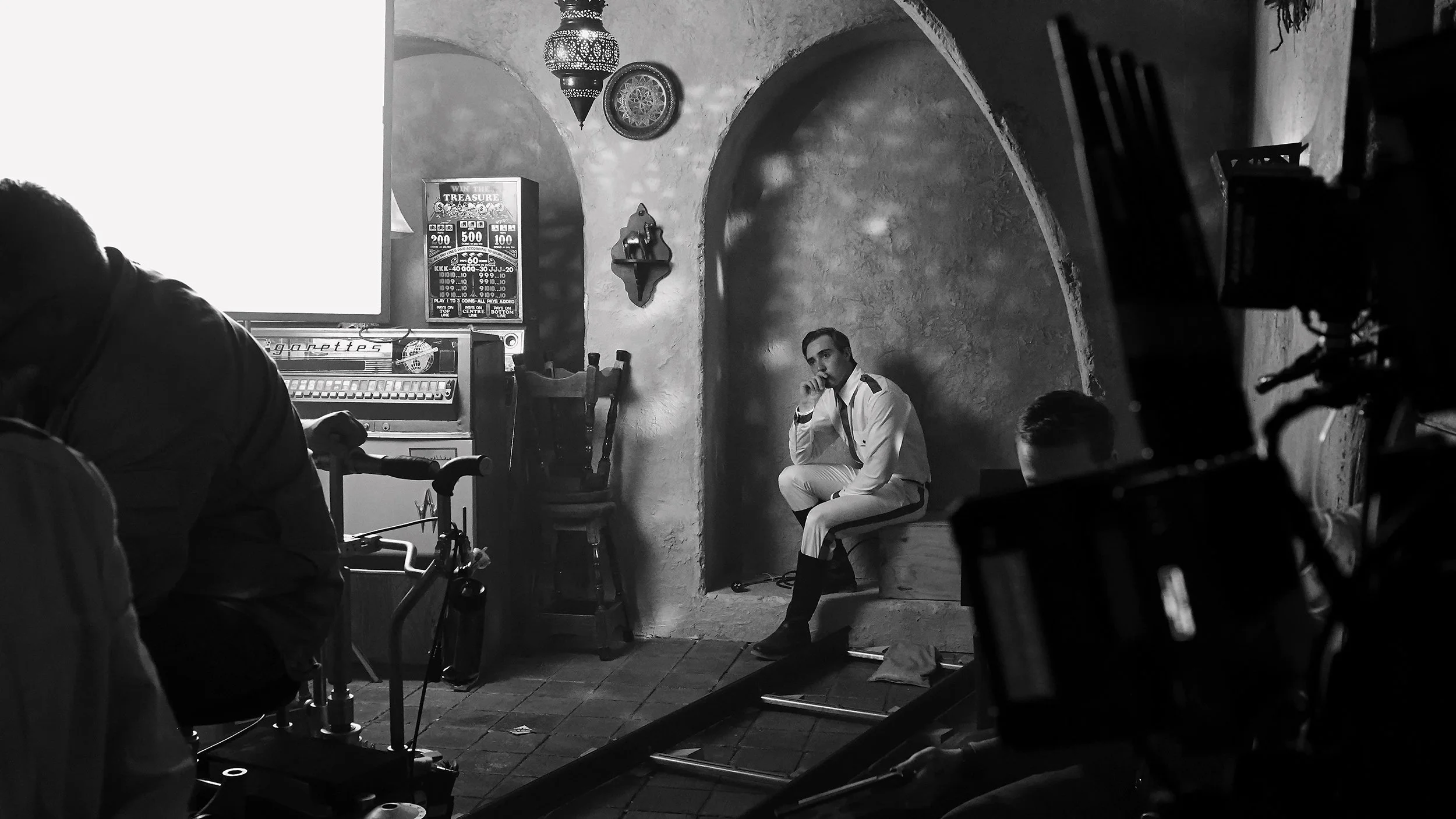 Black and white photo of a man in white clothing sitting on a chair in a rustic brick and plaster room. There are chairs and a pinball machine in the background. The man is looking to the side with his hand near his mouth, and other people and camera