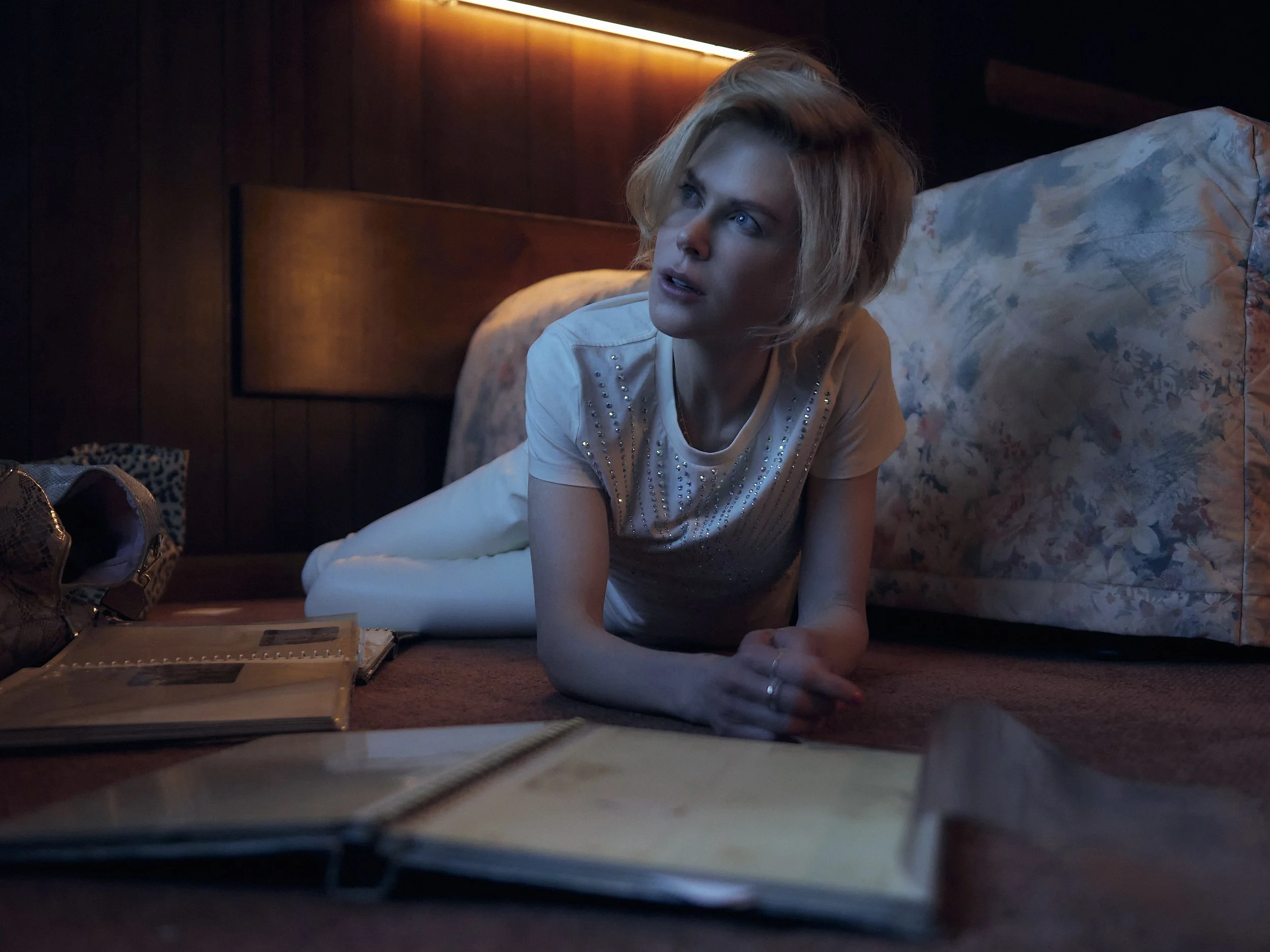 A young woman with blonde hair is lying on the floor of a dimly lit room, propped up on her elbows, looking thoughtfully into the distance. She is wearing a white shirt with embellishments. There is a floral pillow behind her, and paperwork and a not