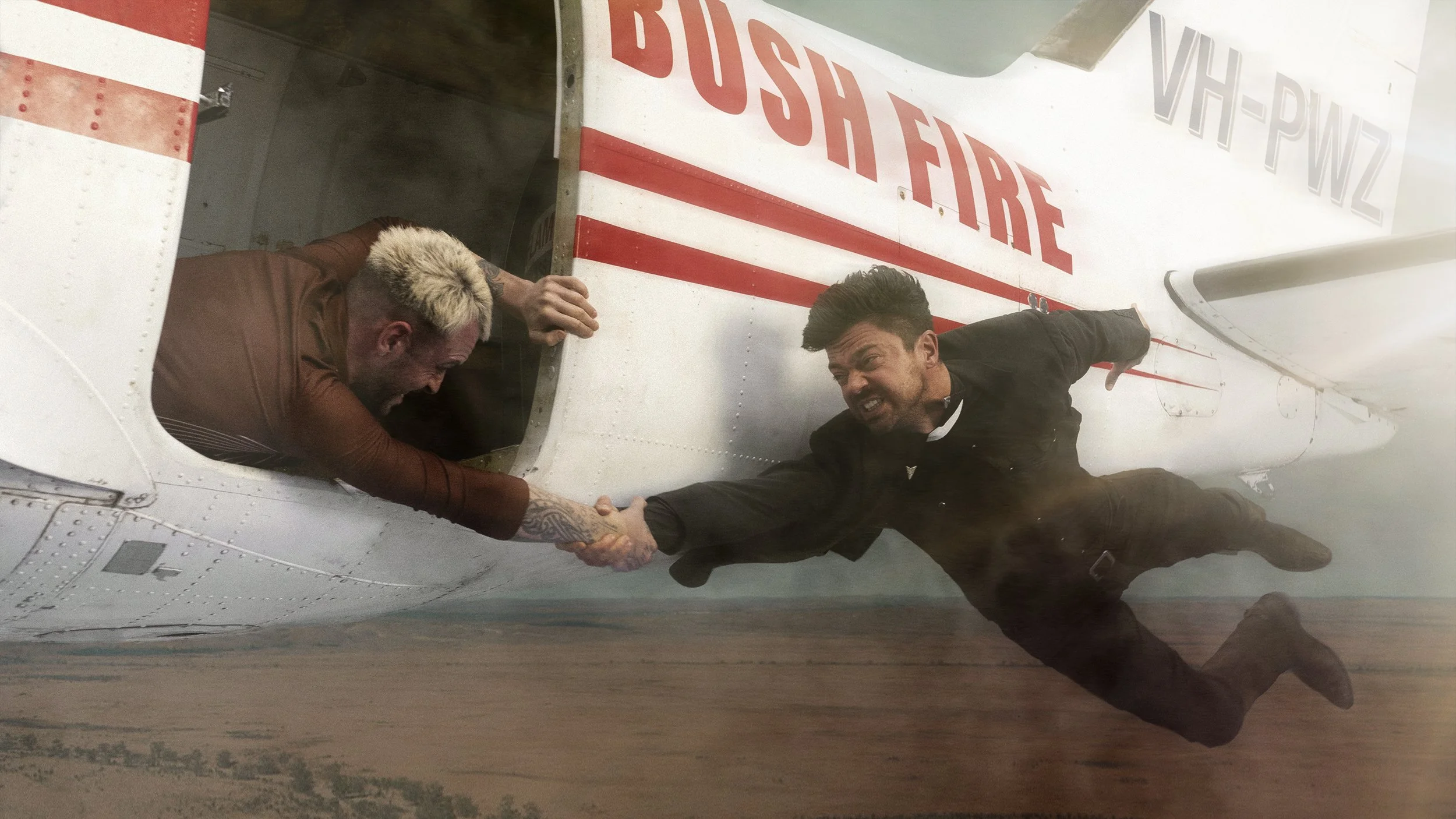 Two men are performing a stunt on an aircraft fuselage marked with 'BUS STOP FIRE'. One man with blonde hair and tattoos on his arm is inside the aircraft, reaching out, while the other man in black is hanging outside, gripping the first man's hand, 