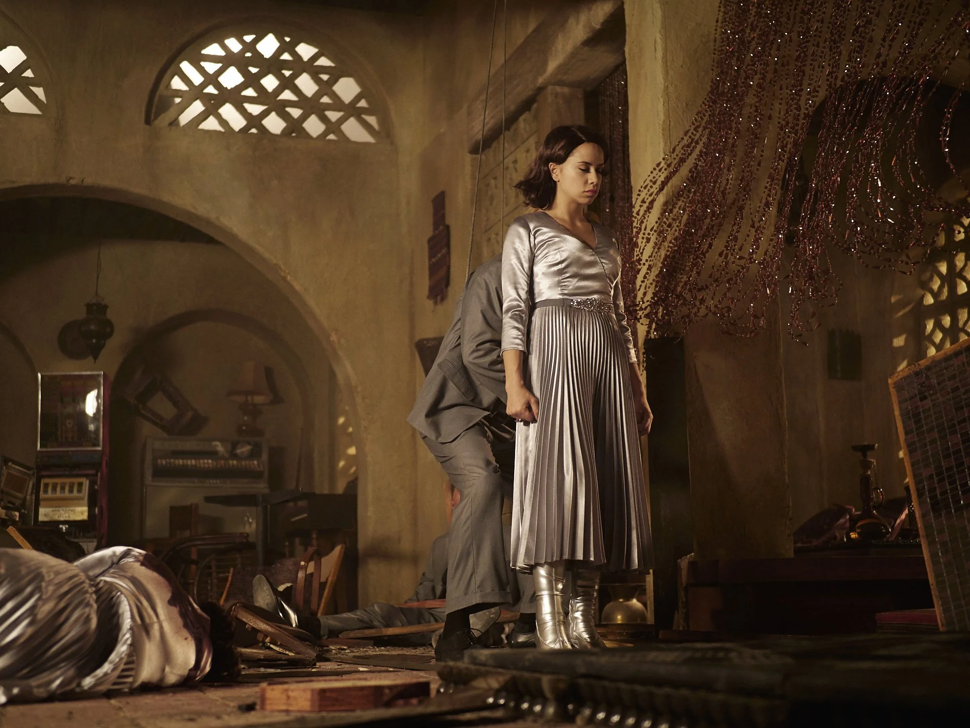 A woman with dark hair and silver dress stands on someone’s back, who is lying on the ground, in a warmly lit, rustic interior with arches and decorative elements.