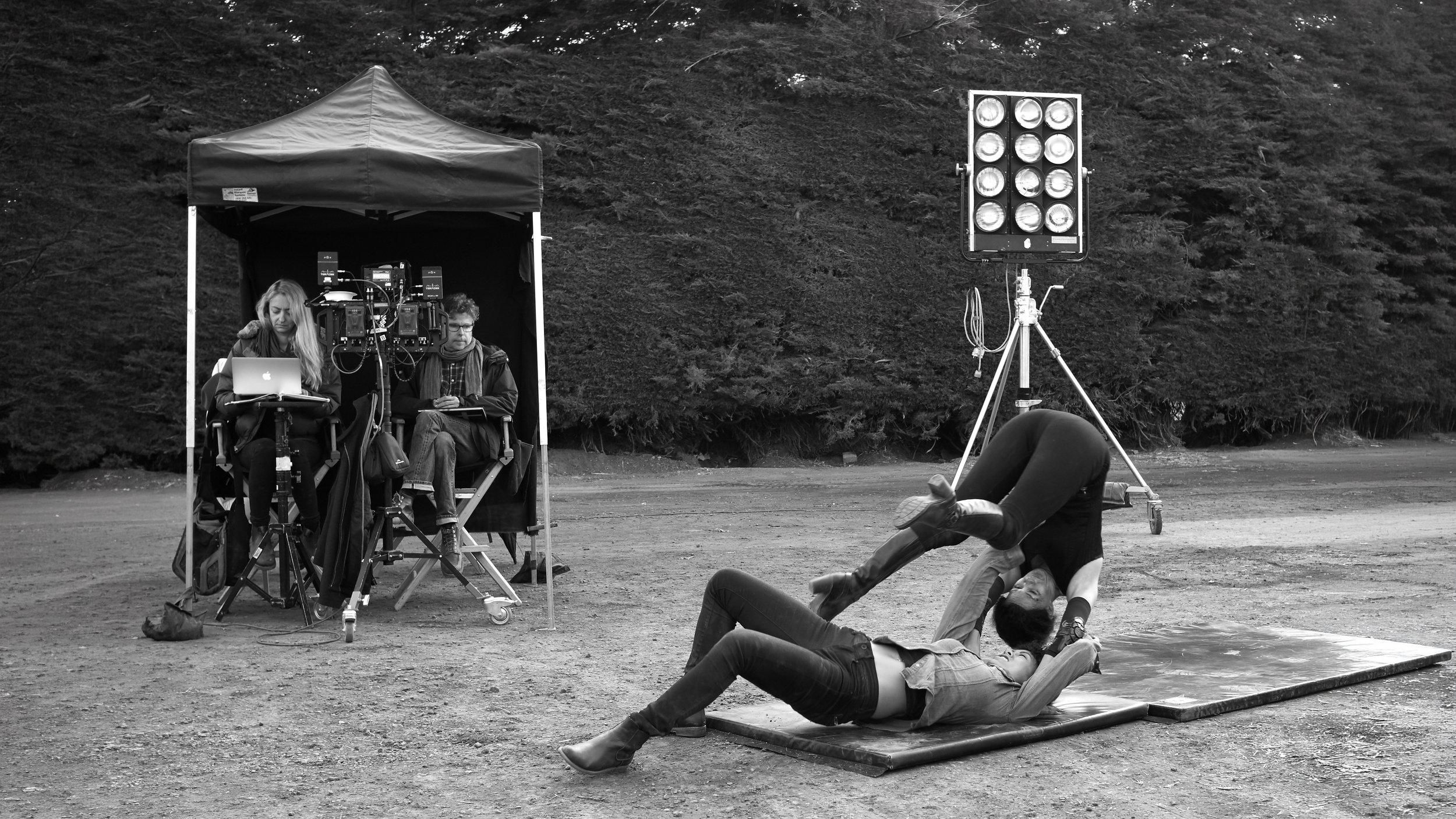 Filming scene outdoors with two people in a tent, one working on a laptop and the other on a camera setup, large light stand nearby, and a person falling on a mat while another person helps them during a stunt.