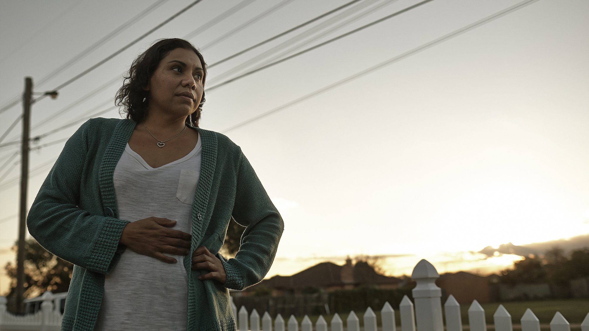 A woman standing outdoors near a white picket fence during sunset, wearing a gray t-shirt and green cardigan, with her hand on her stomach.