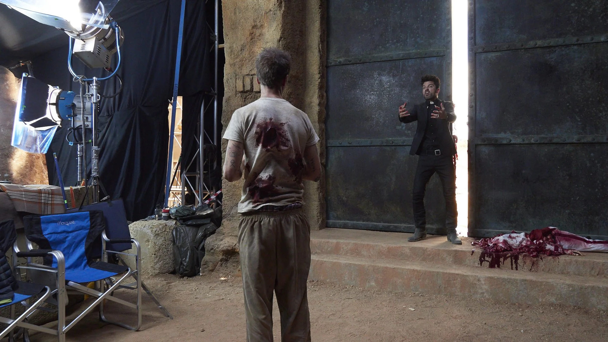 Behind the scenes of a film or TV production, a man with fake bloodstains on his back stands on stone steps, facing another man with a beard who appears to be speaking or directing. There is production equipment, including lighting, nearby, and a lar