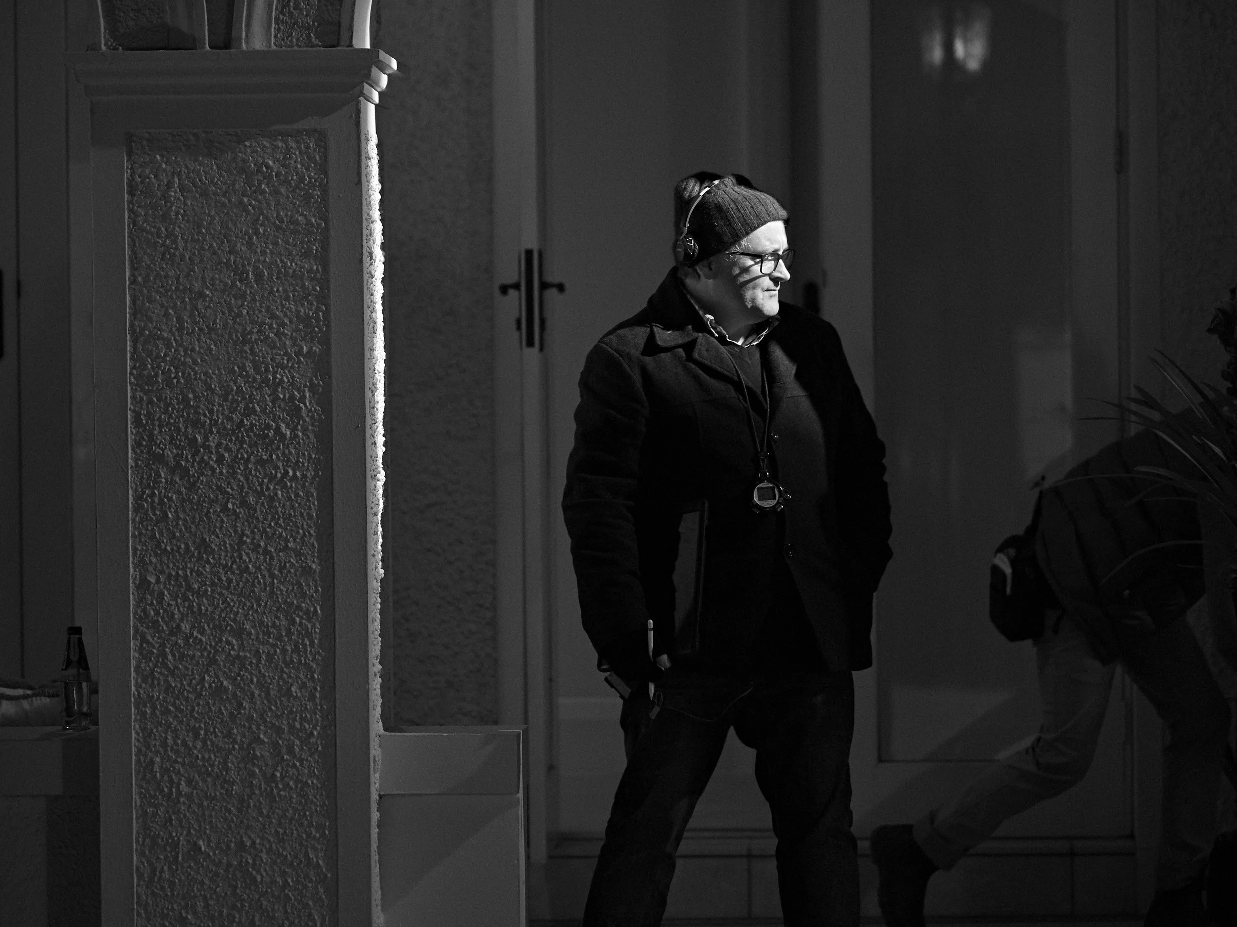 A man wearing glasses, a beanie, and headphones stands inside a building near a textured column. The scene is shown in black and white.