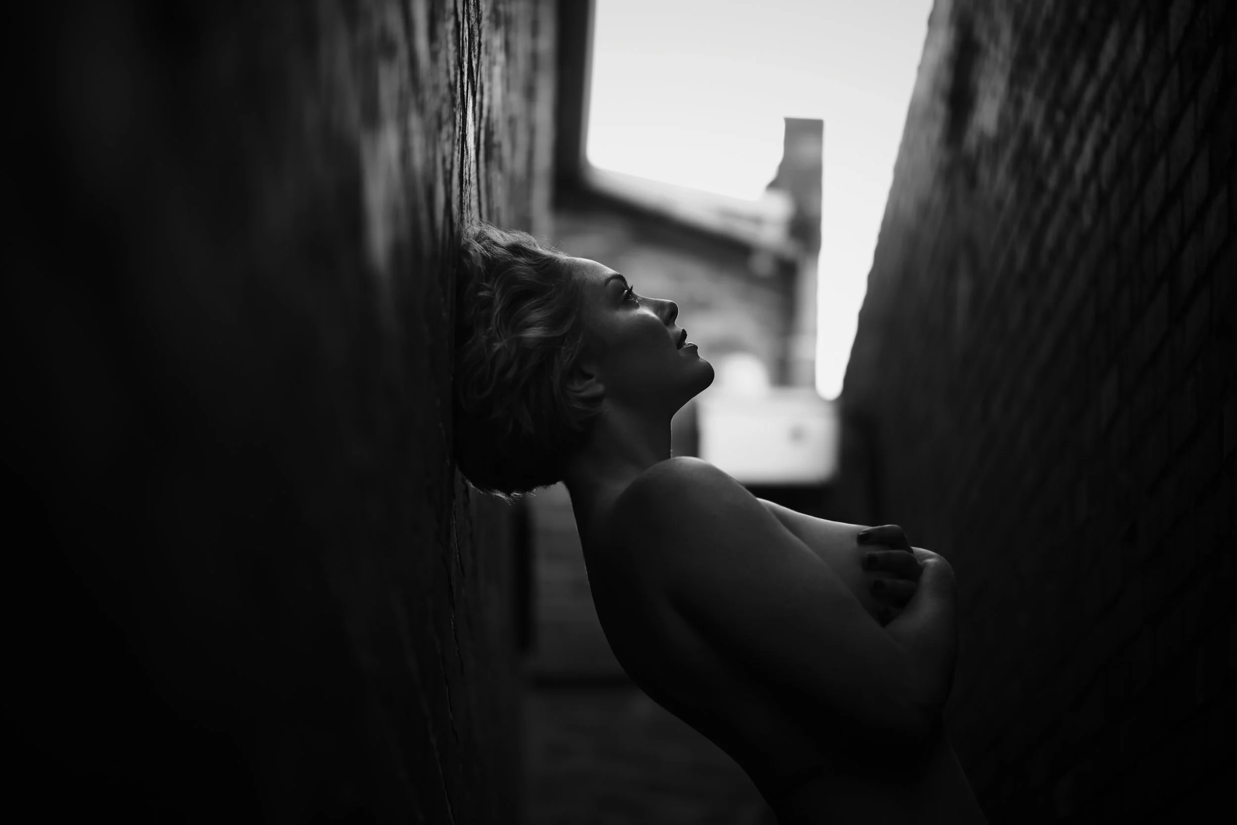 A woman lying down on her back between two brick walls, with her head resting on the ground and looking upward, in black and white.