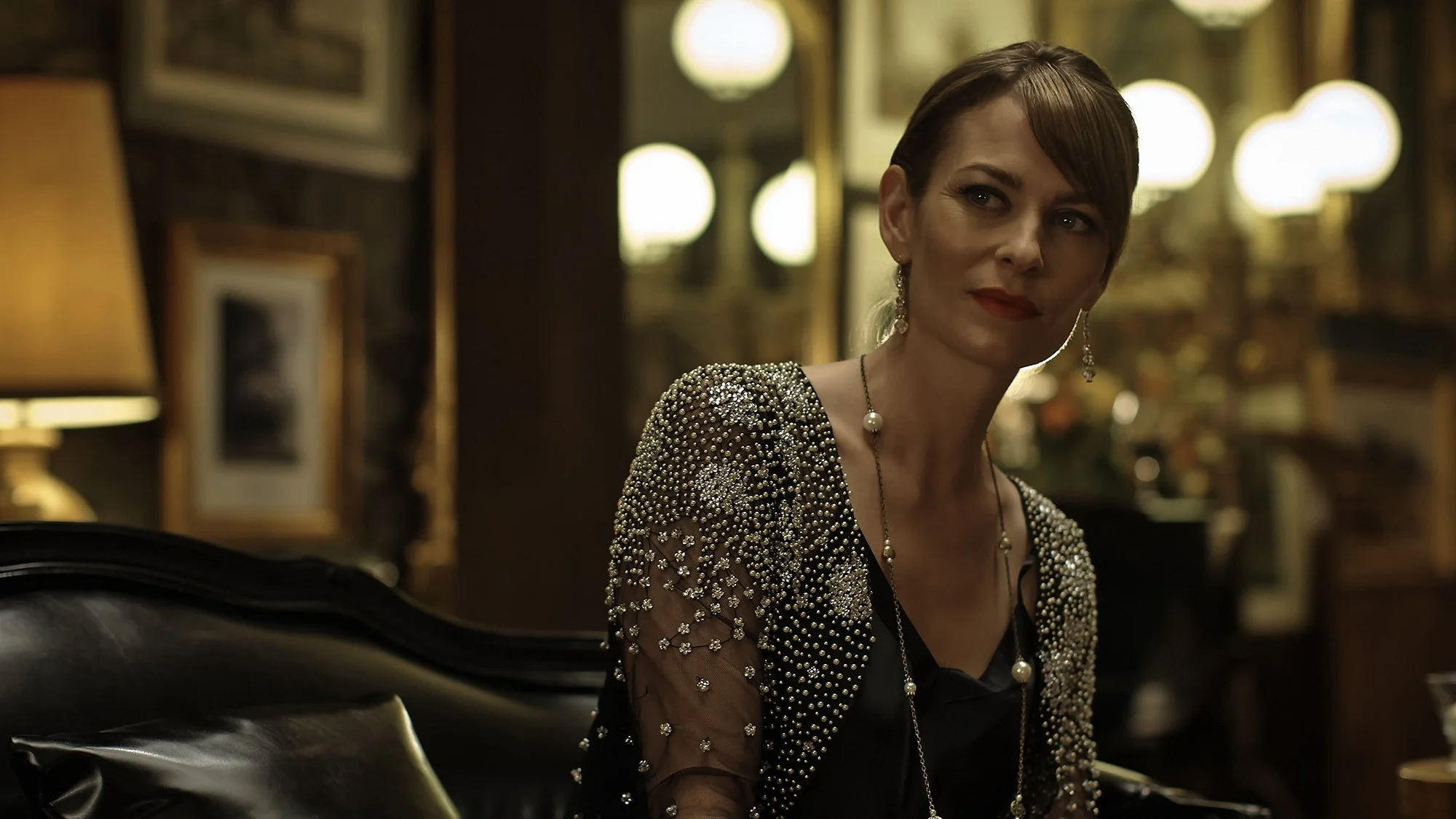 A woman with short brown hair wearing a black dress and a sparkly jacket with pearl embellishments, sitting in a warmly lit room with framed pictures and lamps in the background.