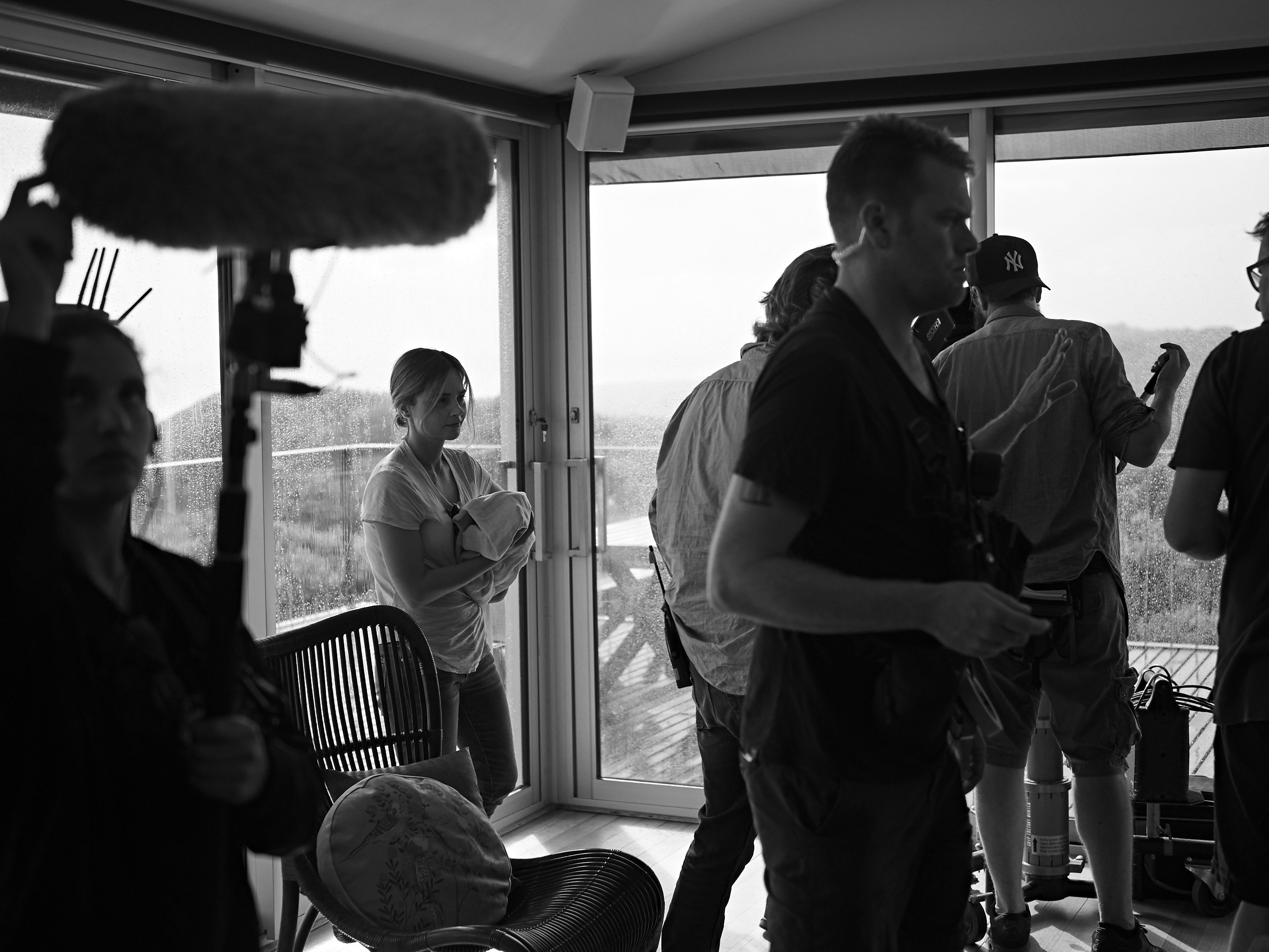 Multiple people gathered indoors, some wearing ear protection and engaged in conversation or activities, with large windows showing an outdoor landscape.