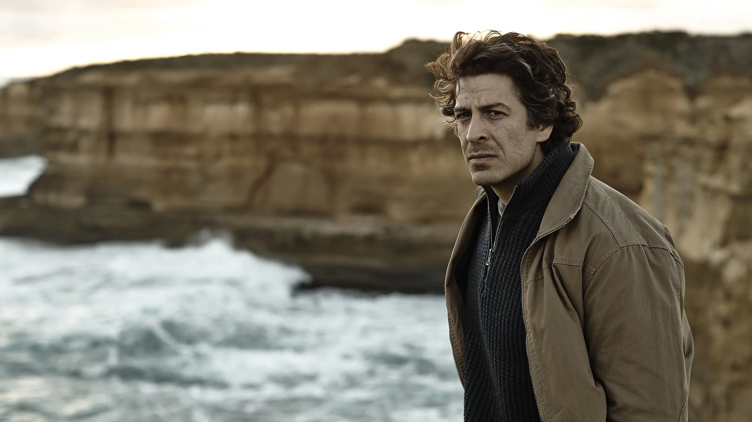 A man with curly dark hair and a serious expression standing outdoors near cliffs and the ocean, wearing a tan jacket and a dark sweater.