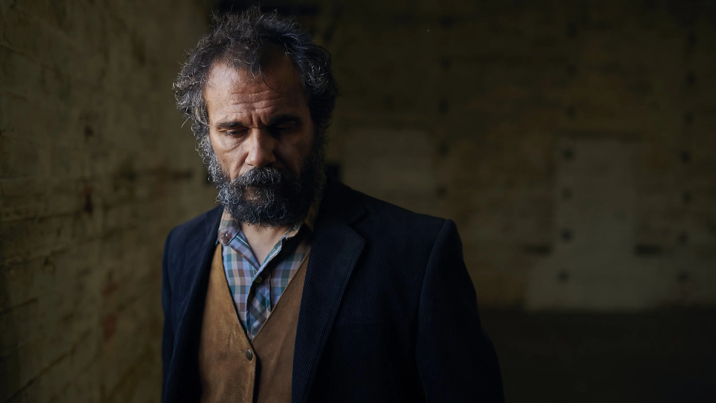 A middle-aged man with a beard and curly hair standing in front of a weathered brick wall, wearing a dark blazer over a plaid shirt and a brown vest, looking down.