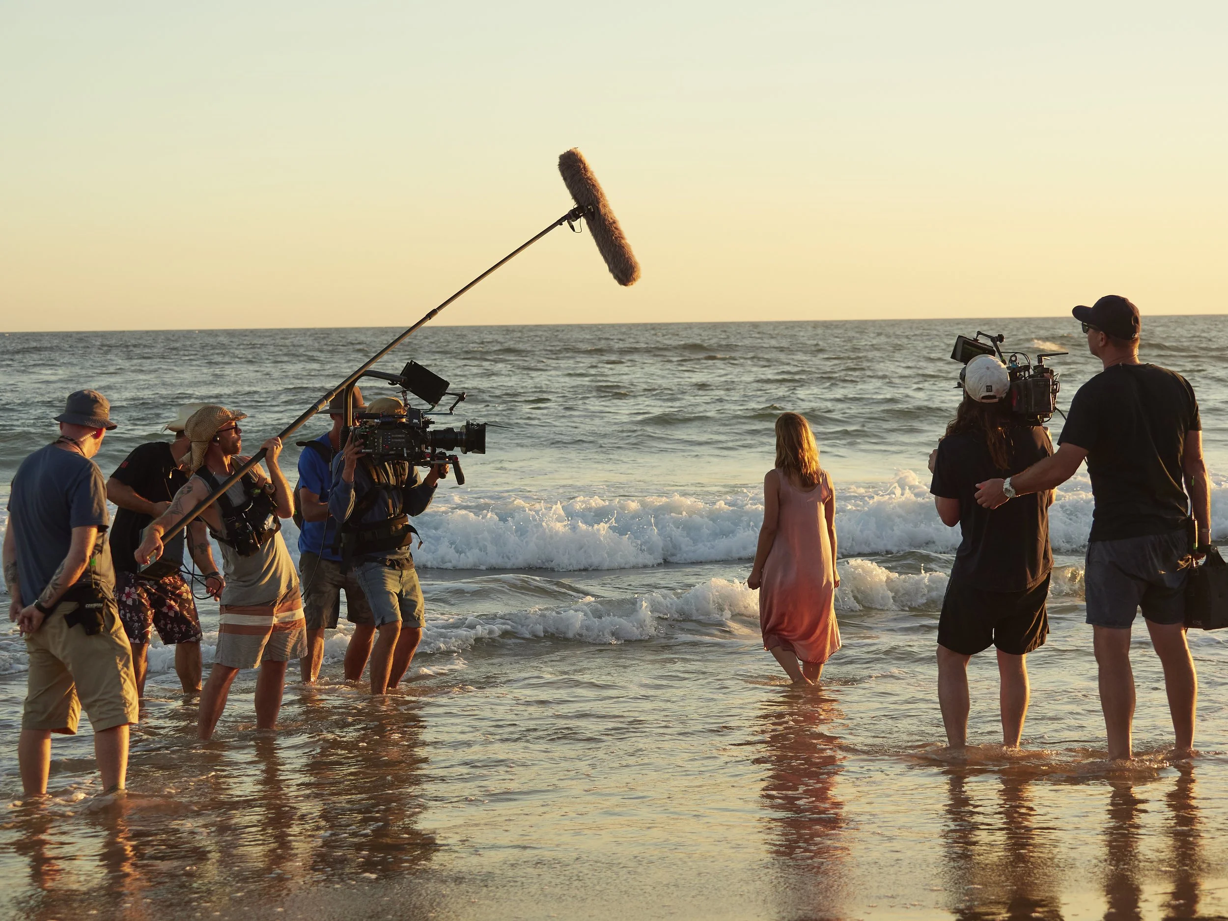 Film crew filming on the beach at sunset, with people walking in the shallow water and a boom microphone overhead.