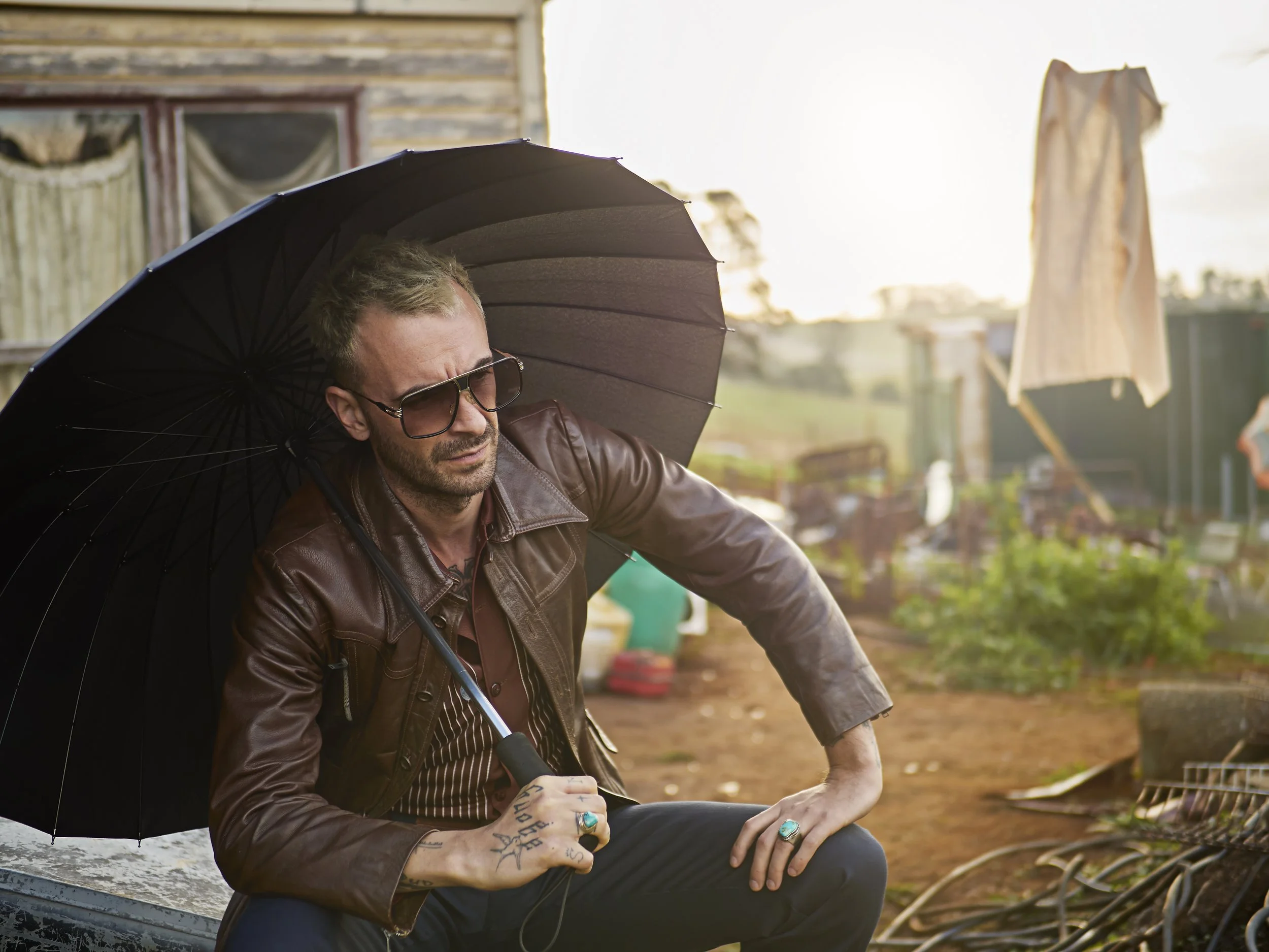 A man with tattoos and rings sitting outdoors with a black umbrella, wearing sunglasses and a brown leather jacket, with laundry hanging on a line behind him and a rustic building in the background.