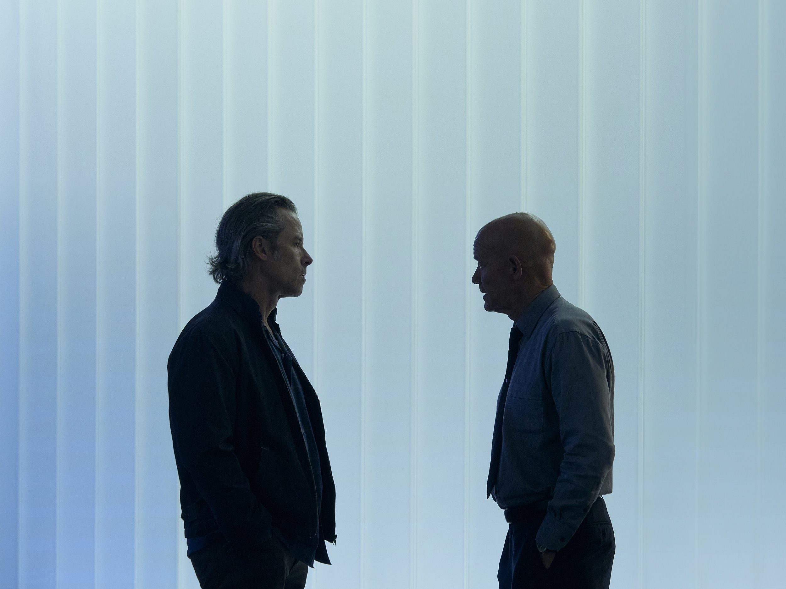 Two men face each other, a silhouette created by the backlighting from a white, vertical-paneled wall.