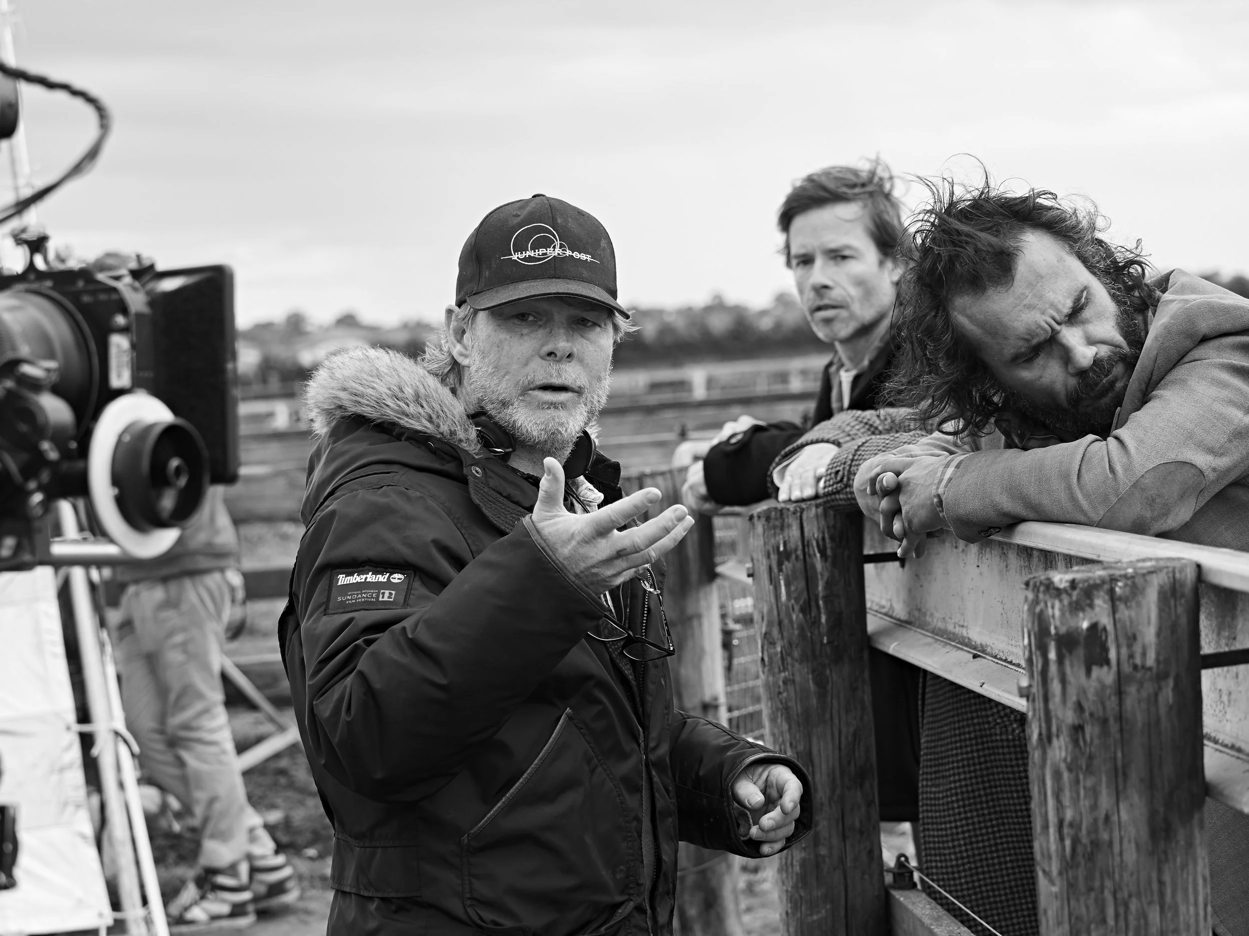 Three men at a film set or outdoor filming location, with one man in the foreground wearing a black jacket and a baseball cap, gesturing while sitting by a wooden railing, and two men behind him looking in his direction, with a film camera in the for