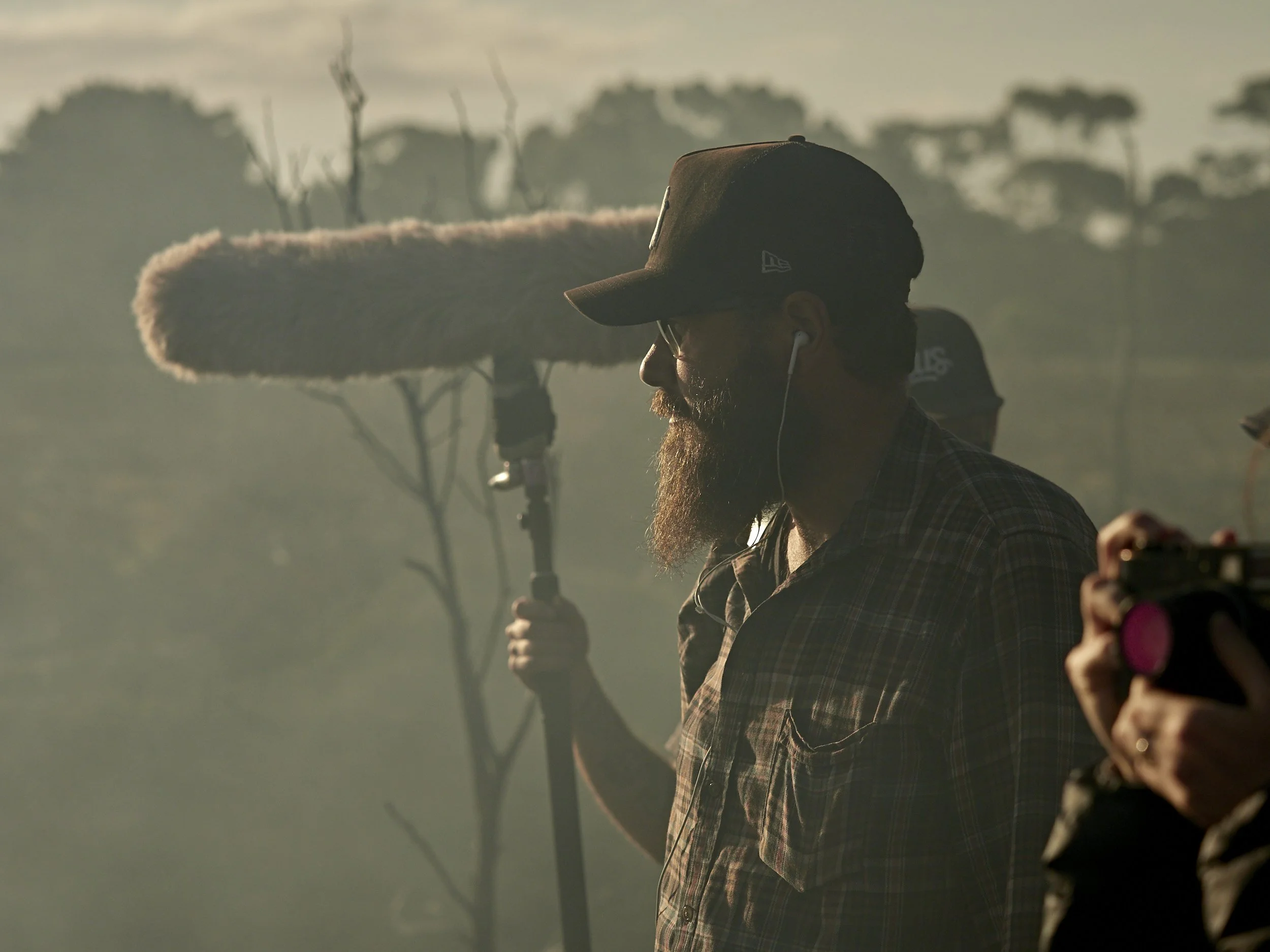 A man with a beard wearing a baseball cap and plaid shirt using a boom microphone at sunrise or sunset in a natural outdoor setting.