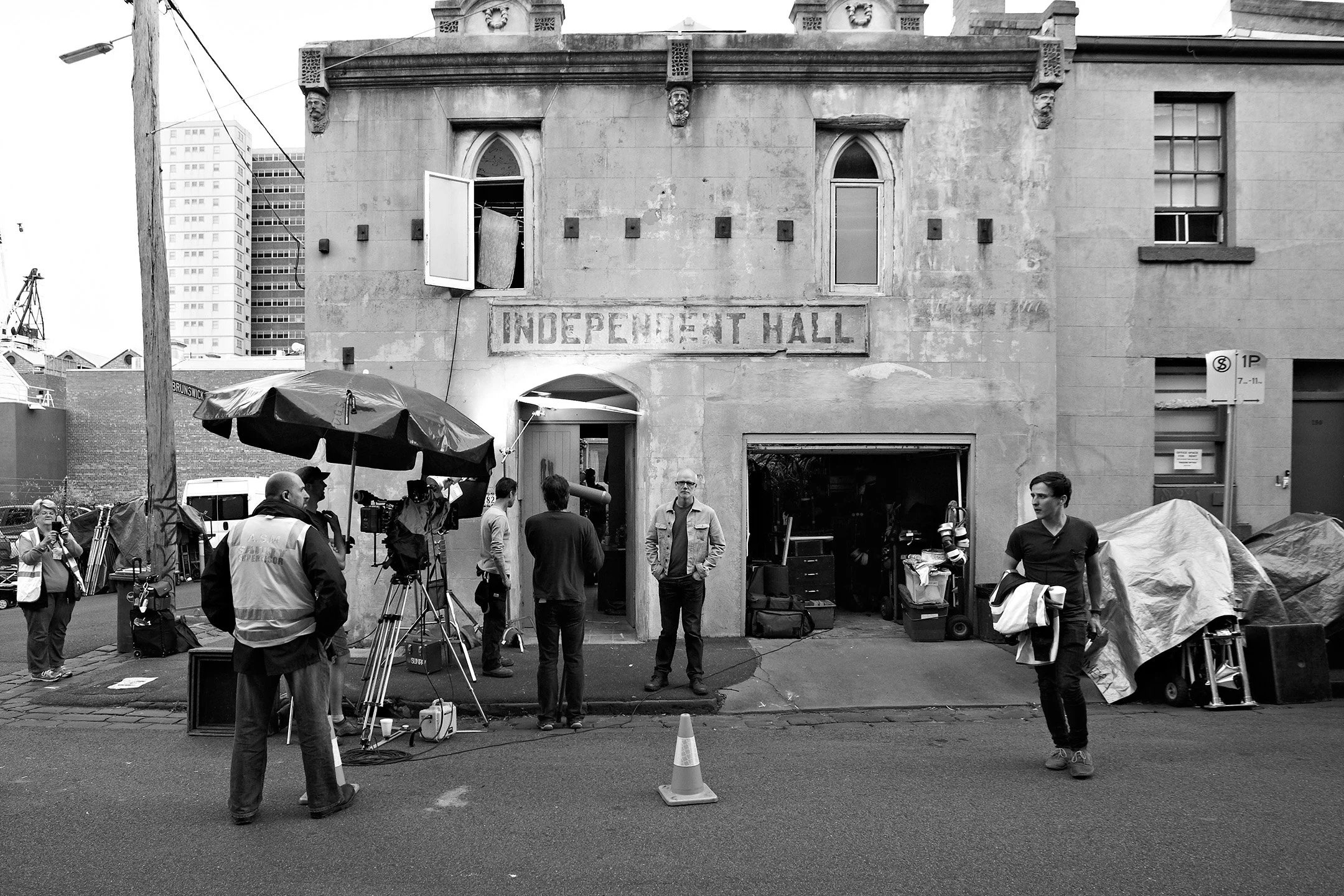 A black-and-white photo of a building labeled 'INDEPENDENT HALL' with people and filming equipment outside. There are camera crews, crew members, and various production gear under an umbrella, with a cityscape of tall buildings in the background.